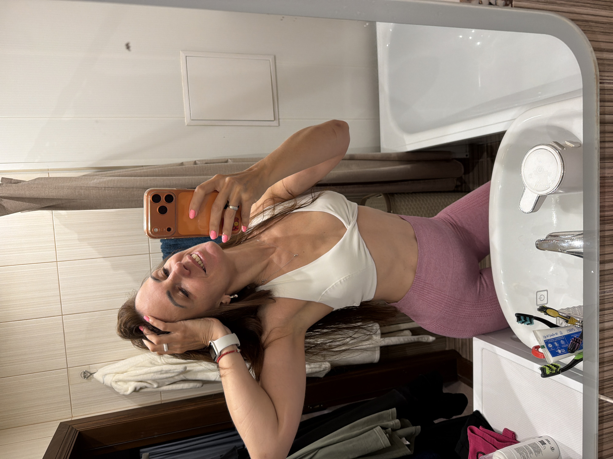 A woman with long brown hair is taking a mirror selfie in a bathroom. She is wearing a white sports bra and pink leggings. Her left hand is on her head, and her right arm is bent, holding an orange smartphone. She is smiling and looking at her reflection. In the background, there is a tiled wall, a bathtub, a sink with toothbrushes and toothpaste, and some clothing hanging on a rack. The bathroom appears to be in a residential setting, possibly in Birobidzhan, Russia.