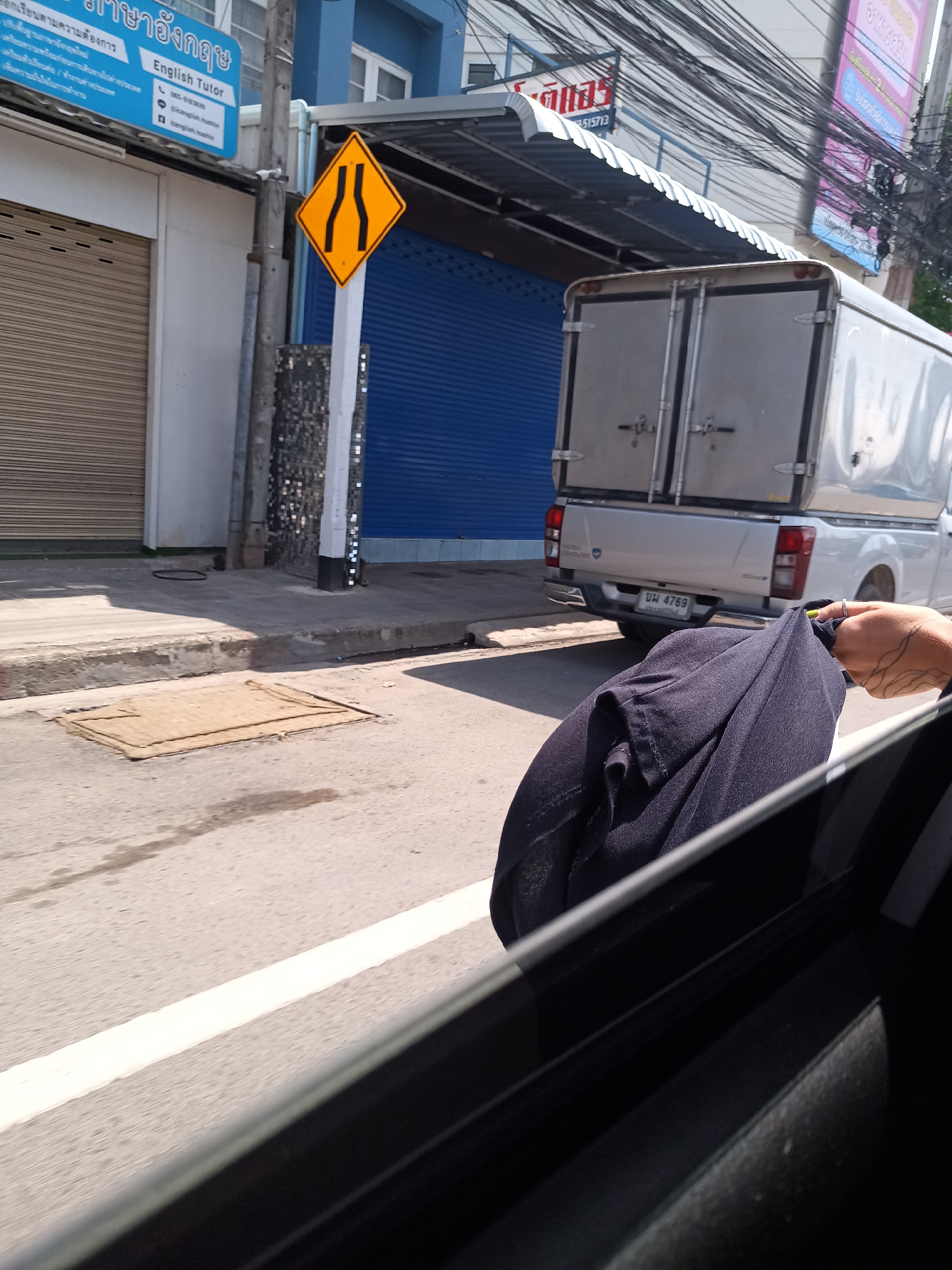 The photograph, captured from inside a vehicle in Hua Hin City Municipality, Thailand, presents a street scene. The view includes a yellow road sign indicating the narrowing of the road, adjacent to a building with a blue sign that reads "English Tutor." A pickup truck with a silver-colored cargo bed is visible on the right side of the frame, its license plate reading "บม 4769." The road is marked with white lines, and a sewer cover is visible on the asphalt. A person's hand and a dark blue garment are in the foreground, partially obscuring the view from inside the car. Overhead power lines are also visible.