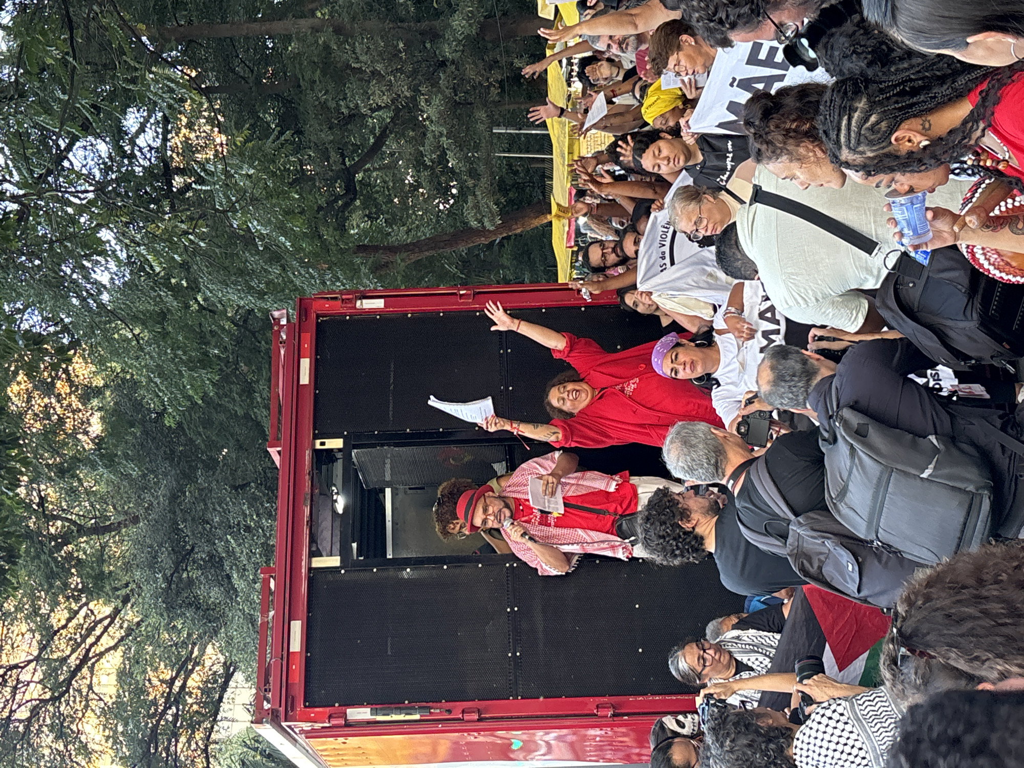 A crowd of people gathered in São Paulo, Brazil. Many of them are concentrated around a red truck, on top of which two women and one man are speaking, one holding a microphone. The crowd is looking at the speakers, many with arms raised. Some are holding up pieces of paper. Many people are wearing black backpacks. The speakers are on a black, flat surface, and wearing red shirts. The trees are visible in the background.