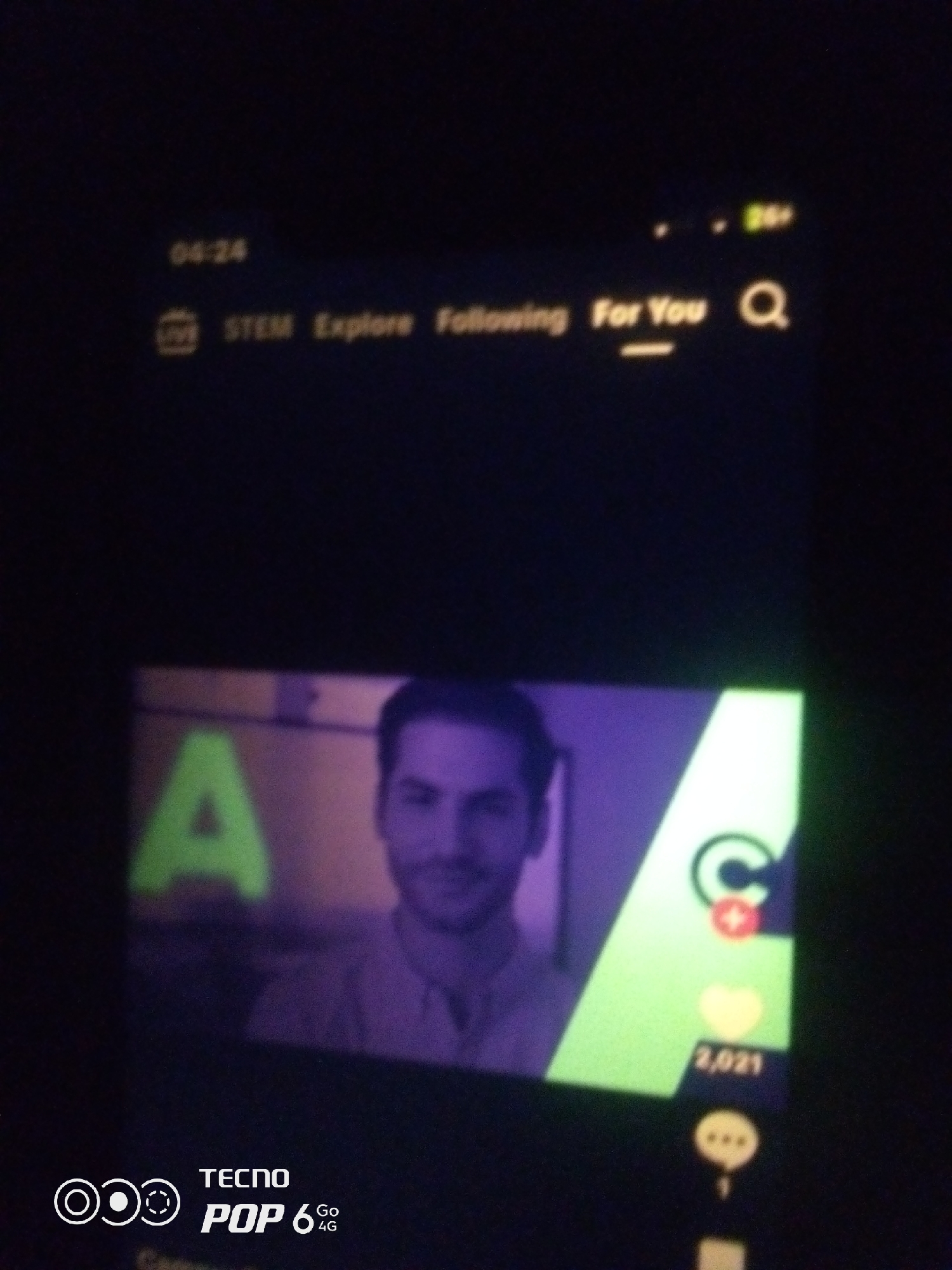 The image displays a smartphone screen showing a social media interface, likely a video-sharing platform. The time displayed at the top left is "04:24". Across the top, there are navigation options: "LIVE", "STEM", "Explore", "Following", and "For You", along with a search icon. The main content of the screen is a video featuring a man with dark hair and a beard. To the left of the man's face, a large, bright green letter "A" is visible. To the right of the man, a bright green area contains icons, including a red plus sign and a yellow heart with the number "2,021" next to it. At the bottom of the screen, a logo for "TECNO POP 6 Go 4G" is displayed. The overall scene is dimly lit, with the screen providing the primary illumination. The image is of low resolution and appears to have been taken in a dark environment, potentially at night. There is no visible indication of the city of Howayi, Nigeria, within the image itself.