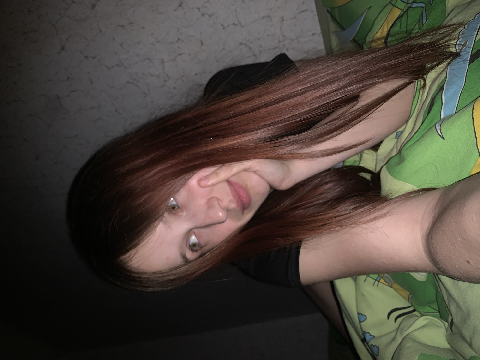 A young woman with long, auburn hair is resting on a green and yellow patterned bedcover. She is wearing a black shirt. Her right hand is touching her face near her ear. The background includes a mottled beige wall. The location appears to be indoors, presumably in a private residence. No specific details indicate this is Фоки, Russia.
