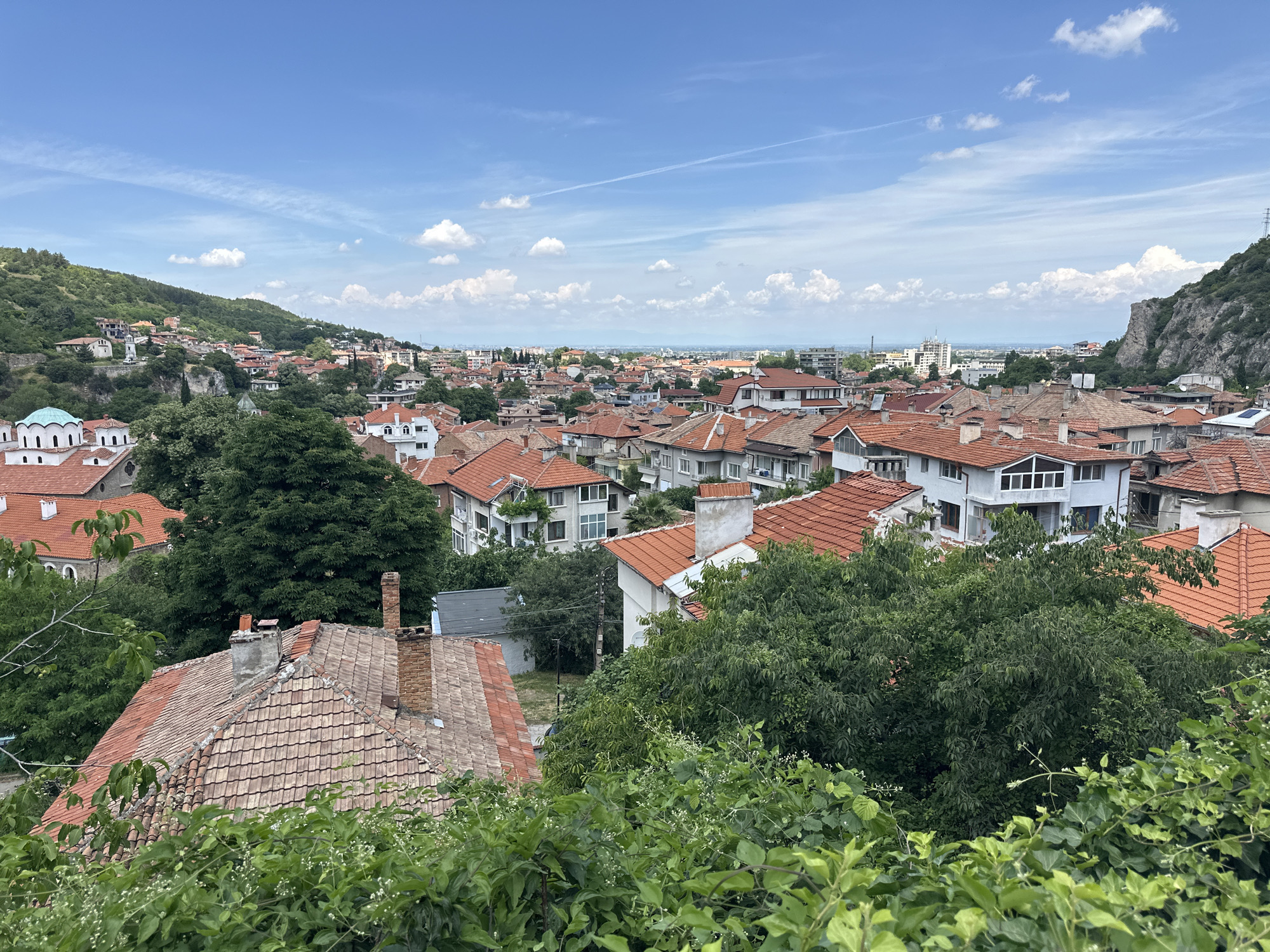 This is a daytime, elevated, wide-angle shot of the city of Asenovgrad, Bulgaria, taken from a high vantage point, likely a hill or a tall building. The foreground is dominated by lush green foliage, obscuring the lower portion of the view.
Beyond the vegetation, the scene unfolds into a panorama of red-tiled rooftops, characteristic of Mediterranean and Balkan architecture. Numerous residential buildings, varying in height and style, are densely packed together, creating a textured urban landscape. Many of these buildings feature white or light-colored facades.
In the mid-ground, a prominent church with a distinctive turquoise dome and arched windows stands out, adding a significant architectural landmark to the city. Further into the distance, the urban sprawl extends, with a mix of older and more modern buildings, including some taller, multi-story structures and apartment blocks.
The backdrop of Asenovgrad is characterized by rolling green hills that recede into the distance. On the right side of the image, a rugged, rocky cliff face rises steeply, forming a dramatic natural element within the cityscape. The sky above is a vibrant blue, dotted with scattered white clouds and a faint contrail from an aircraft. The overall impression is a warm, sunny day in a densely populated city nestled amidst natural beauty.
