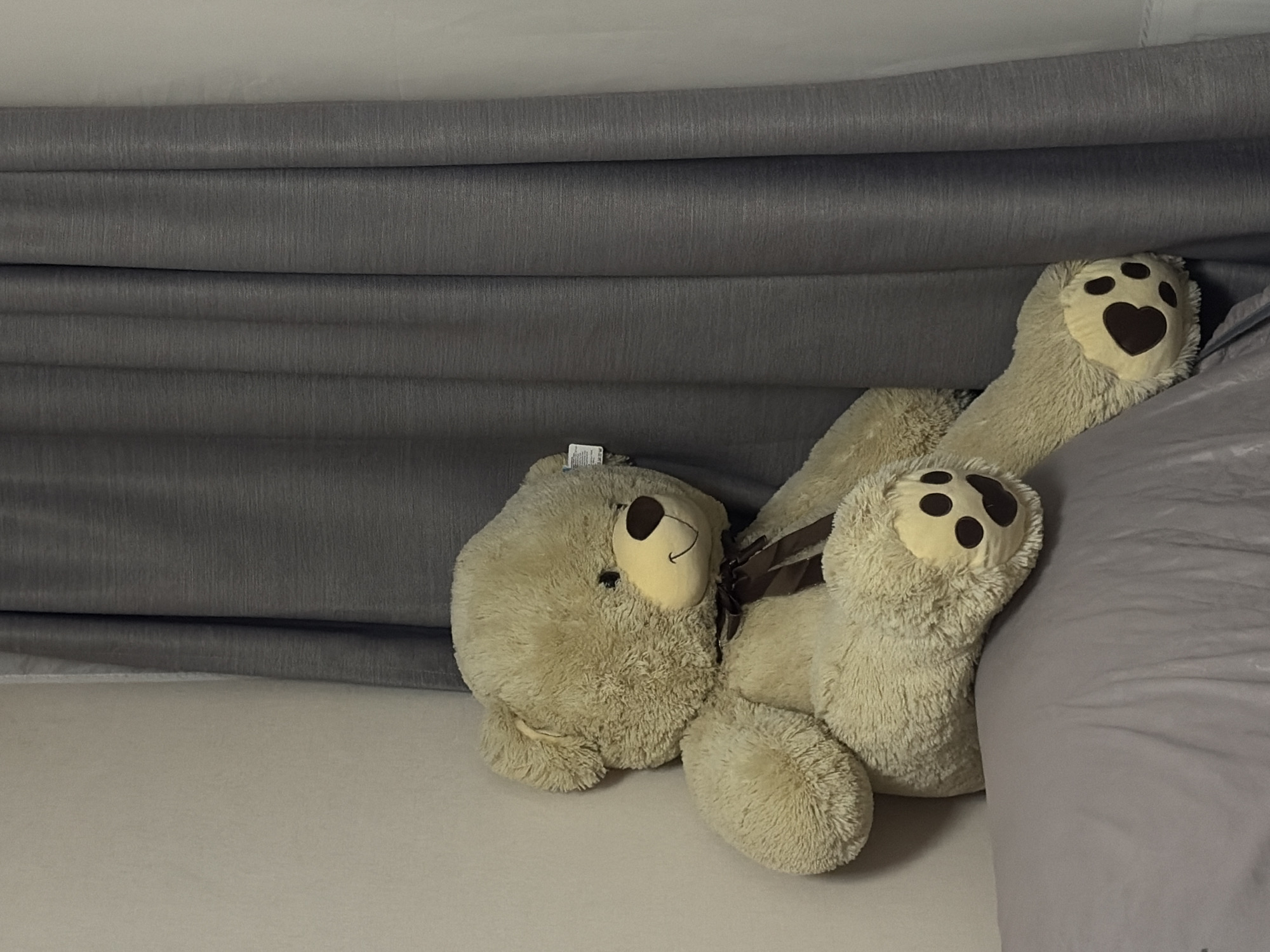 A stuffed bear with heart-shaped paw pads is lying on a bed. The bear has light brown fur and a brown ribbon. The bed has a grey sheet and is positioned against a grey curtain. The image location is unknown and there is no verifiable connection to Krasnodar, Russia.