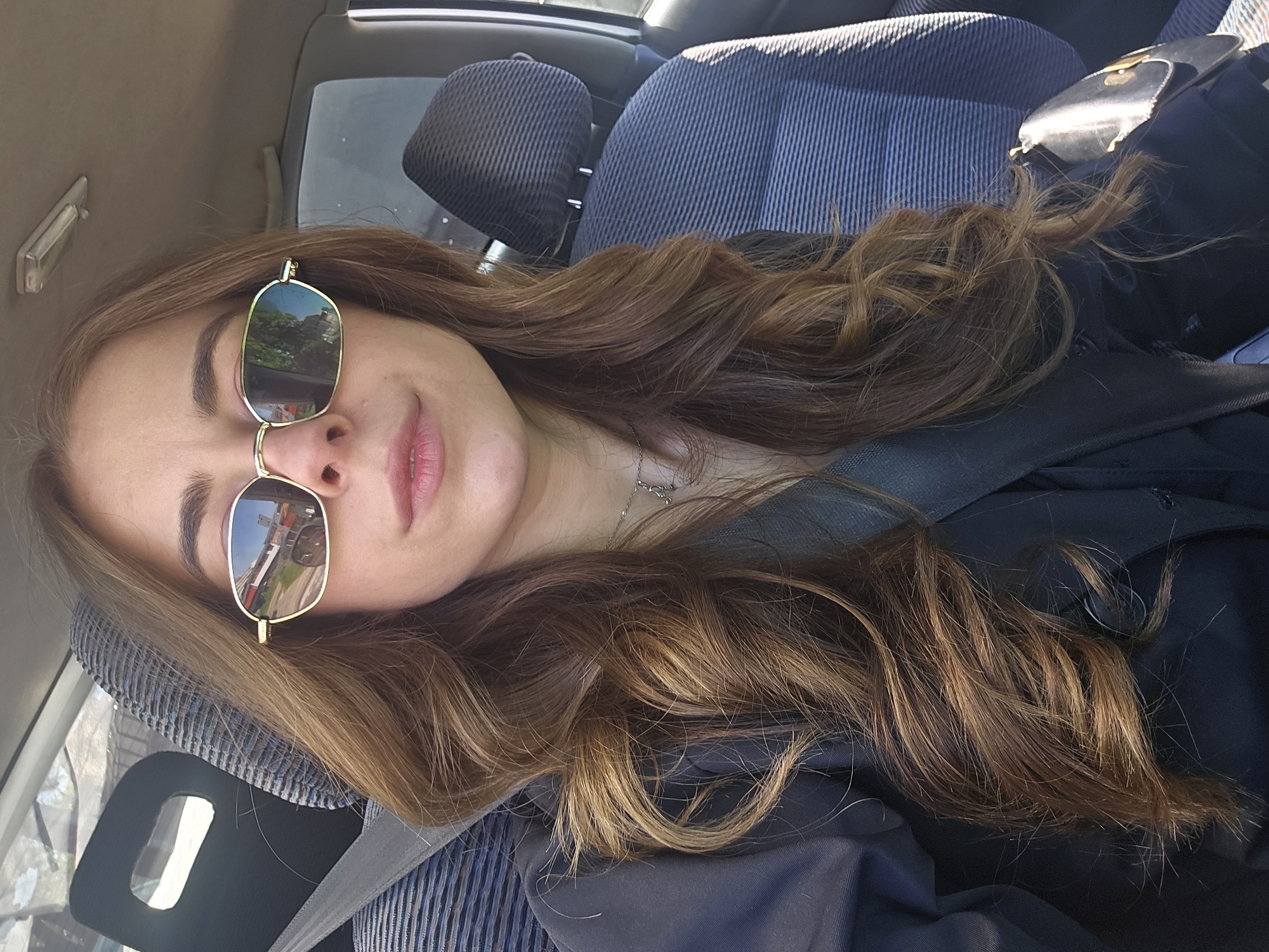 A woman with shoulder-length wavy brown hair is sitting in a car wearing sunglasses. She has light skin and a chain necklace is visible. The car's interior consists of a blue striped seat, a black seat belt, and a black jacket. The reflection in her sunglasses shows a cityscape, including trees and buildings. The location, Plovdiv, Bulgaria cannot be identified in the image.