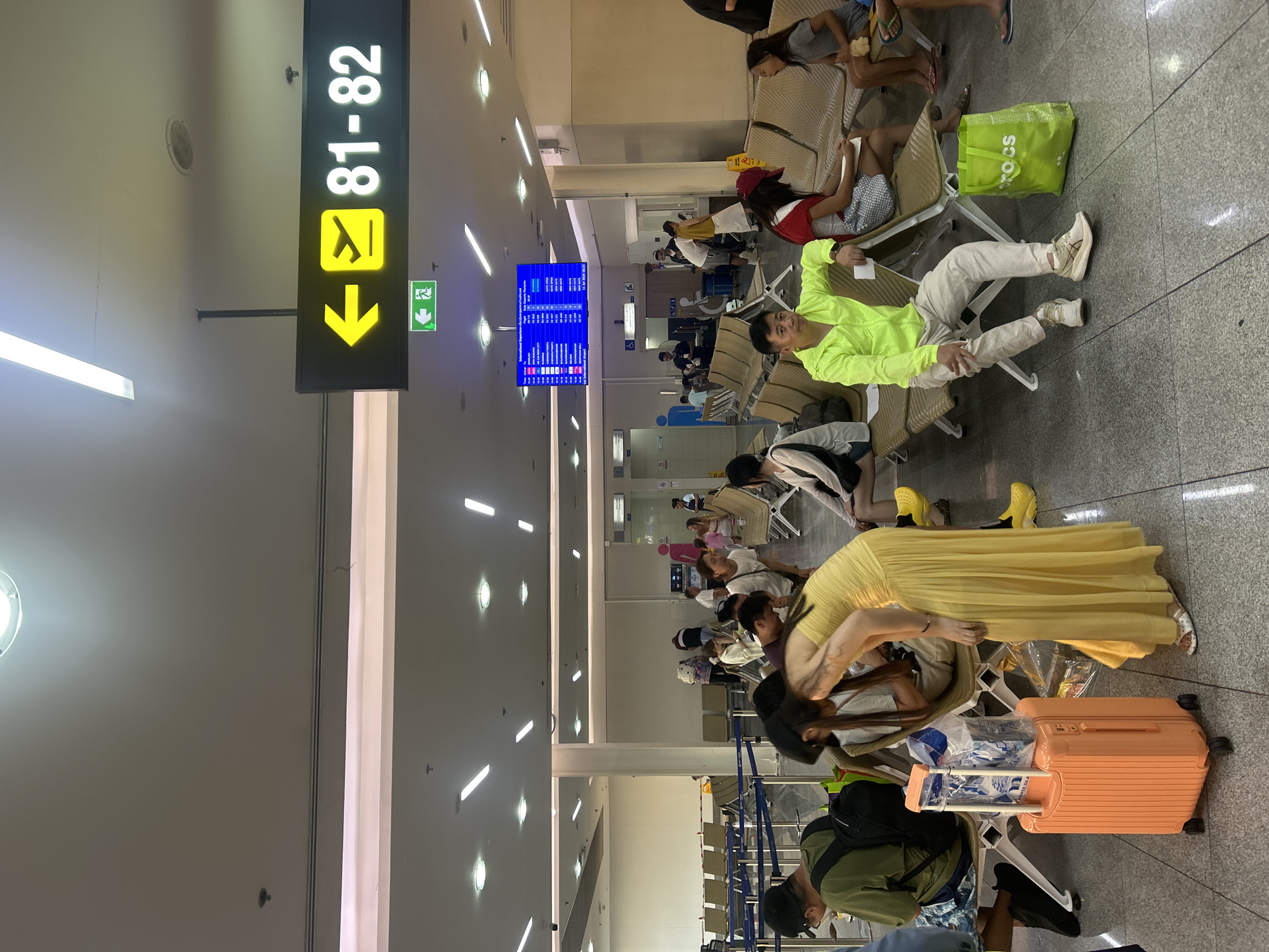 The image depicts an indoor scene, likely an airport departure hall in Mai Khao, Thailand. A group of people are seated on rows of empty chairs, awaiting their flights. The hall is well-lit with fluorescent lighting, and a prominent illuminated sign indicates "81-82" with a directional arrow and an airplane icon, suggesting a gate or boarding area. To the left of this sign, a green illuminated emergency exit sign is visible. Further into the hall, a large digital display screen shows flight information. An orange suitcase is positioned in the foreground next to a woman in a yellow dress. Other passengers are scattered throughout the seating area, some with luggage. The floor is tiled. The atmosphere appears to be one of waiting.