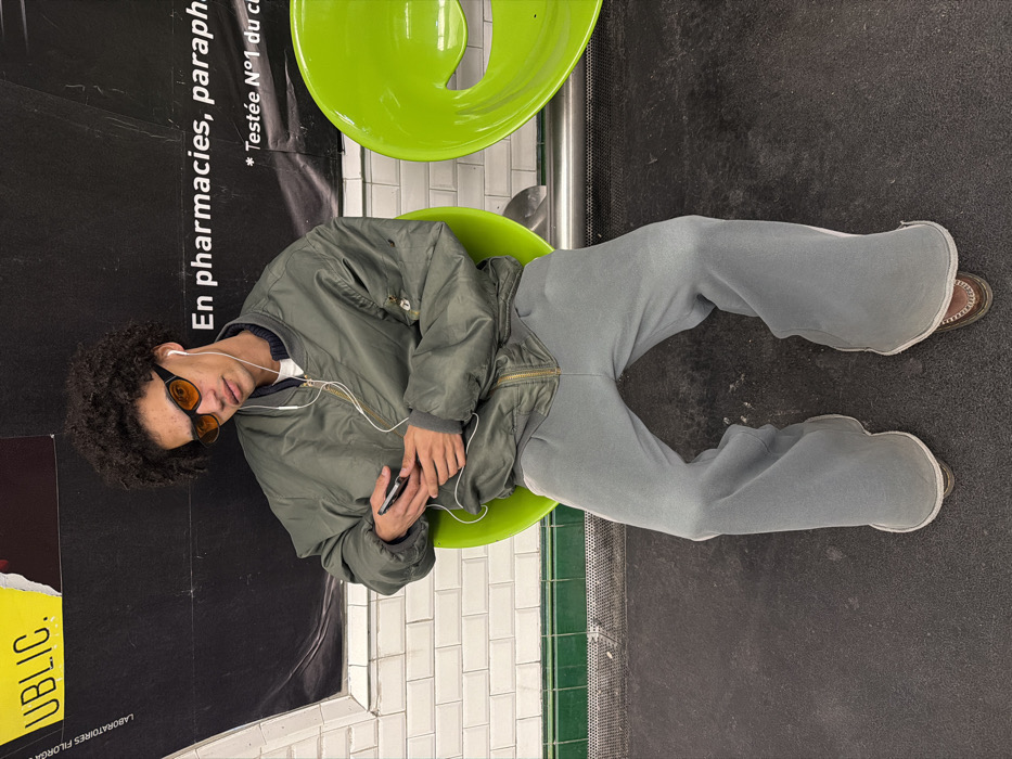 A young individual with dark, curly hair and light-brown skin tone is seated on a lime green ergonomic chair, looking directly at the viewer. They are wearing orange-tinted sunglasses with dark frames and white wired earphones. The individual is dressed in an oversized olive green bomber jacket with a gold zipper and ribbed collar, a white undershirt, and baggy light grey sweatpants. Brown shoes are visible at the bottom of the pant legs. Both hands are clasped around a dark-cased smartphone. To the right, an identical unoccupied lime green chair is visible. The background features a lower section of white tiled wall with a dark green horizontal trim. Above the tiles, a black wall panel displays white French text, including "En pharmacies, parapharmacie" and a torn yellow poster fragment with the word "PUBLIC." The floor is dark grey. The setting is consistent with a public transit station, specifically a Paris Metro station platform or corridor due to the distinctive tiling and French signage.