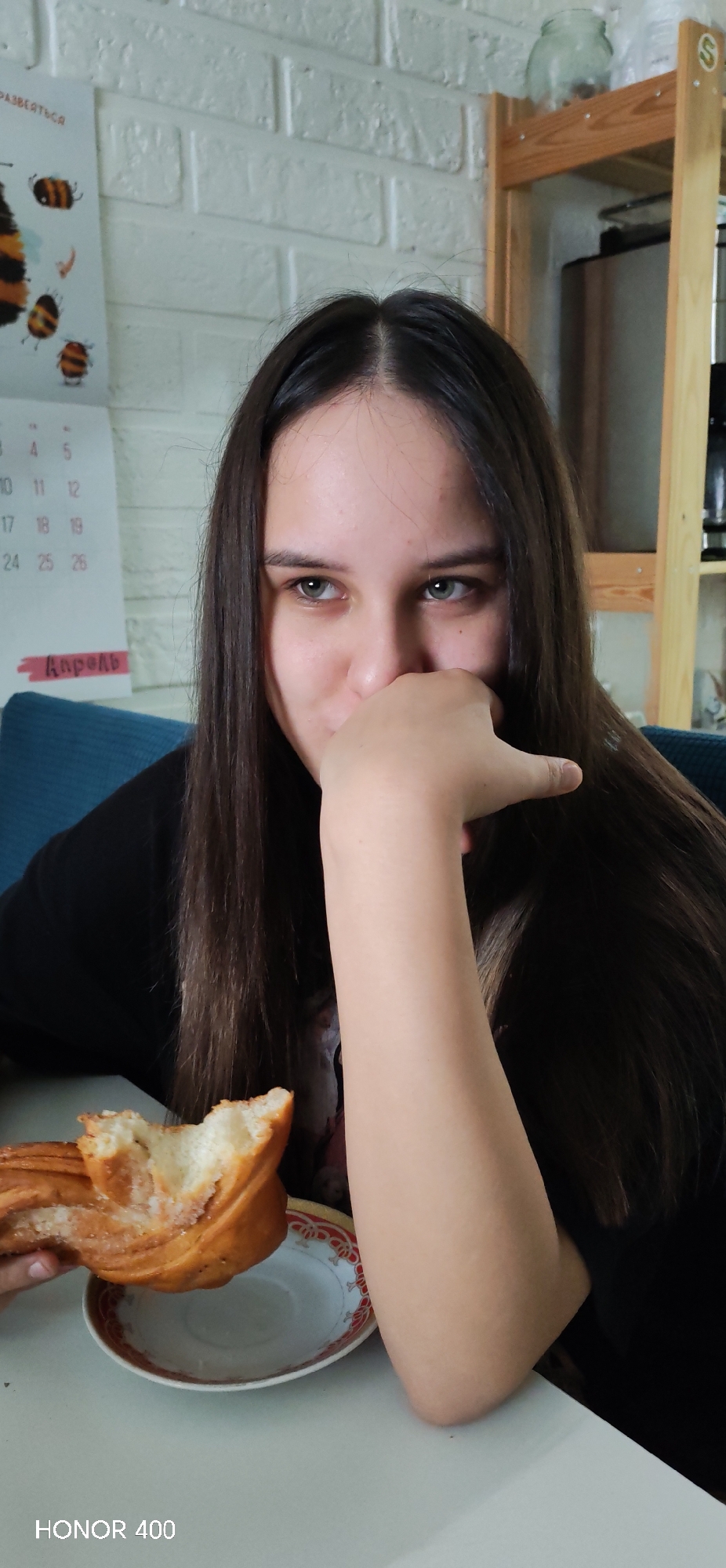 A young woman with long, dark hair parted in the middle is seated at a white table indoors, captured in a close-up, half-body shot. She wears a black top and has her left hand raised to partially cover her mouth, while her light-colored eyes gaze to her right with a subtle expression that hints at amusement or shyness. In front of her on the table is a partially eaten pastry, resembling a sweet bun with a sugary topping, resting on a small white plate with a decorative red and gold rim.
The background features a white wall with a brick-like pattern. A calendar hangs on the wall, displaying Cyrillic text that includes "РАЗВЕЯТЬСЯ" at the top and "Апрель" (April) at the bottom, along with several dates like 3, 4, 5, 10, 11, 12, 17, 18, 19, 24, 25, 26. To the upper right, a wooden shelf unit is visible, holding a clear jar and a dark appliance, with an 'S' logo affixed to one of the shelves. A blue cushion or part of a sofa is seen behind her left shoulder. The scene appears to be set during the daytime in a domestic environment, possibly a kitchen or dining area in Noviy, Russia. The image has a "HONOR 400" watermark in the bottom-left corner.