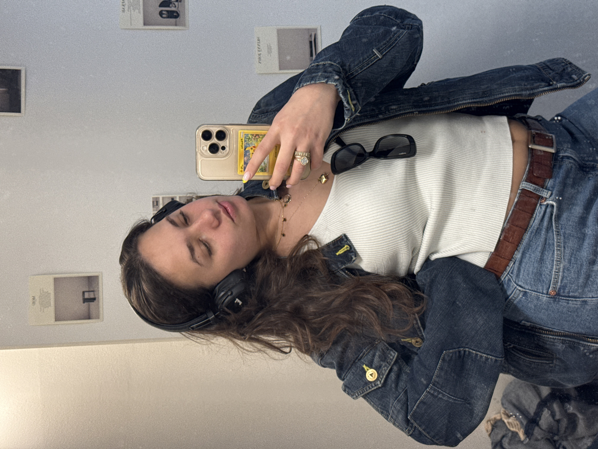 A woman takes a mirror selfie in what appears to be an indoor space, likely a store fitting room or dressing area, due to the presence of framed images on the wall. The location cannot be conclusively identified as Milan, Italy from the image alone. The woman is wearing headphones, a white ribbed top, a denim jacket, and denim jeans with a brown belt. She is holding a cell phone in her right hand, revealing a yellow phone case with a Pokémon card displayed on the back. Rings adorn her fingers, and she wears a necklace. Sunglasses rest on top of her top. She has long brown hair and her eyes are closed. The environment is illuminated with a bright, neutral light.