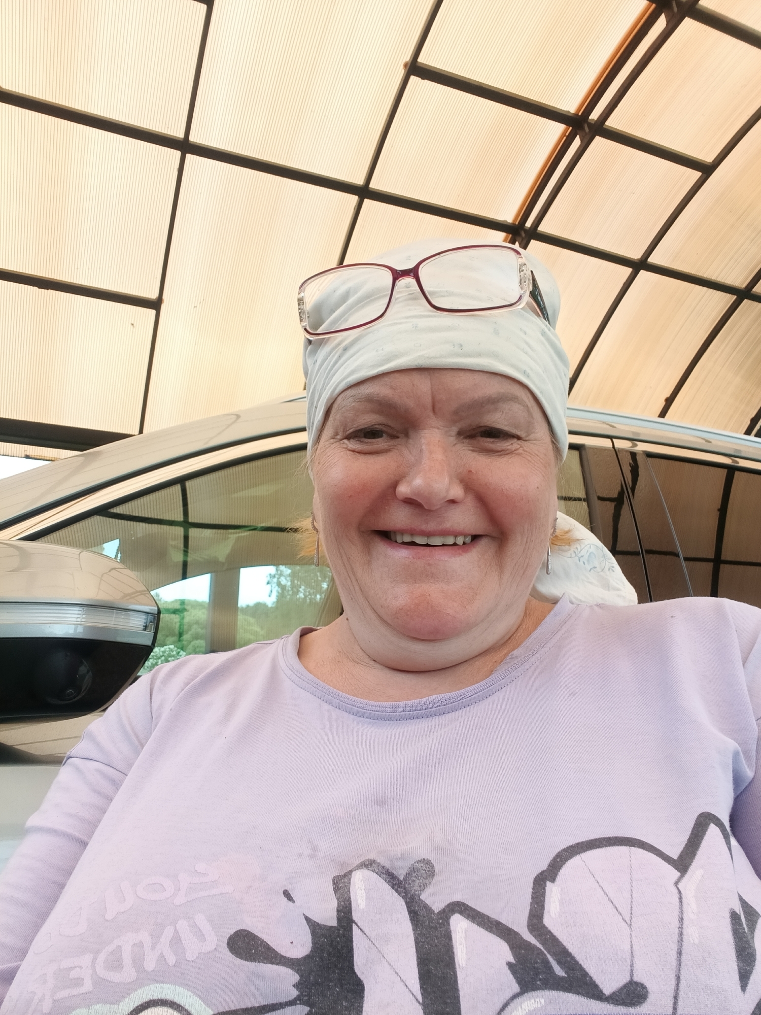 A woman with fair skin, wearing a white headscarf and glasses atop it, is smiling at the camera. She is wearing a light purple t-shirt with a dark graphic design. Behind her is a light-colored car, partially visible, and a structure with a grid-like ceiling. The location is possibly in Красный Маяк, Russia.