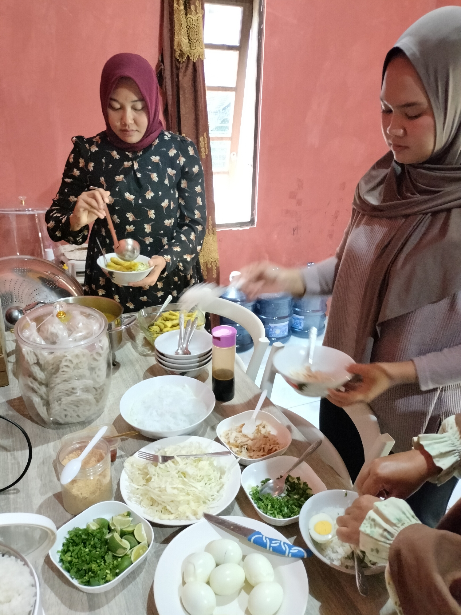 In Angsana, Indonesia, a scene unfolds in what appears to be a residential setting, possibly a home kitchen or dining area, with a pink-painted wall as the backdrop and a window with a dark brown curtain. Two women, both wearing hijabs, are engaged in preparing food. The woman on the left, in a dark floral patterned shirt and a maroon hijab, is holding a ladle over a bowl, scooping out a light-colored broth. In front of her is a large glass jar filled with stacked, round, light-colored crackers. To her right, a table is laden with various food items. These include a pot with a yellow substance, bowls of shredded white cabbage, chopped green onions and lime wedges, and a container of what looks like seasoned breadcrumbs. There are also plates with hard-boiled eggs and bowls of rice, one of which has a sliced hard-boiled egg and some shredded chicken. The woman on the right, wearing a grey hijab and a ribbed sweater, is in the process of serving food into a bowl, holding a spoon and a small plate. Another person's hands, wearing a light-colored sleeve with ruffles, are visible in the lower right corner, also engaged in food preparation, possibly adding egg to a bowl of rice. Several bottles of water are stacked in the background. The atmosphere suggests a communal meal preparation.