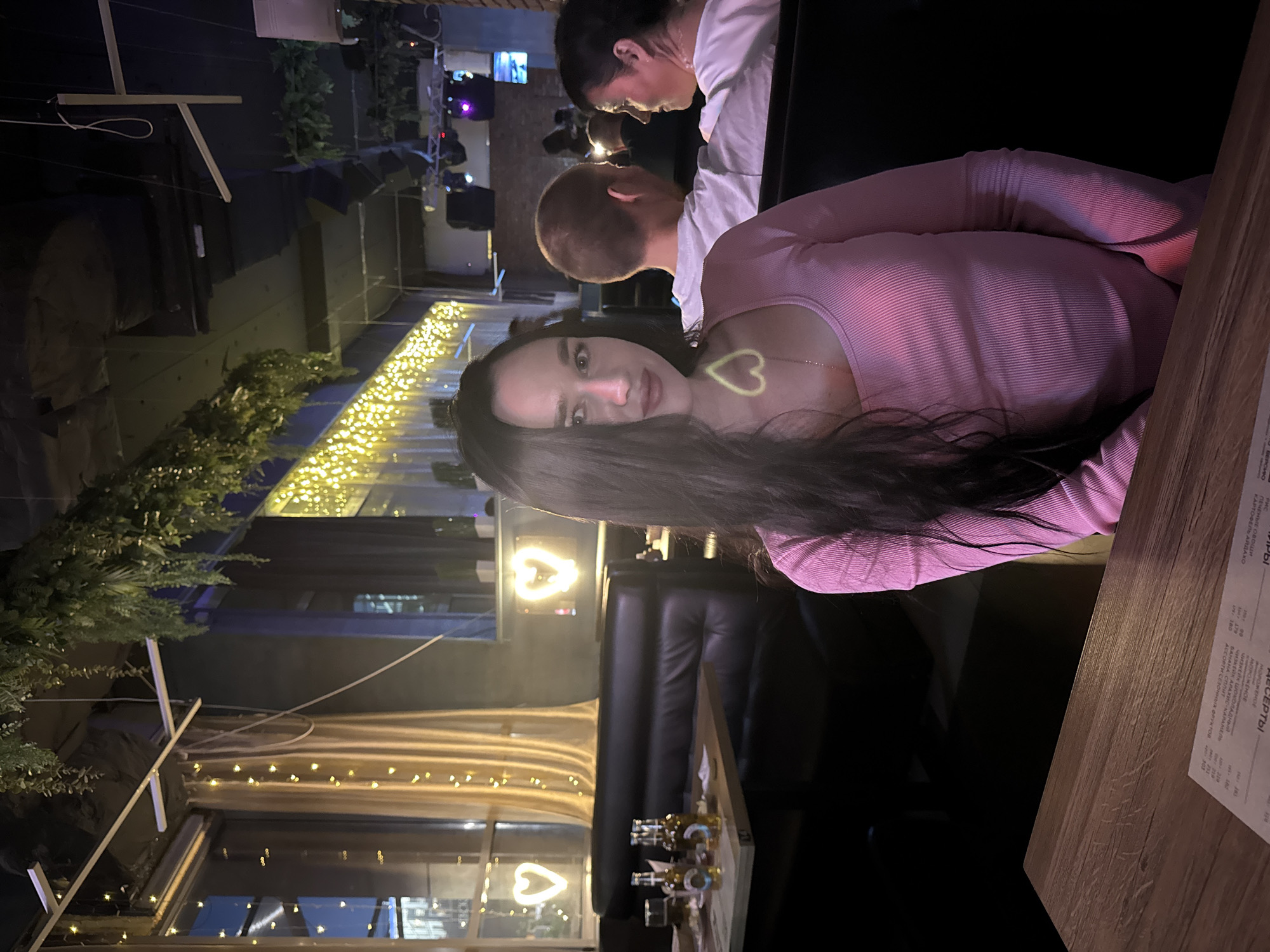 In an indoor establishment, a young woman with long dark hair is seated at a table, facing the camera. She is wearing a pink long-sleeved top. In the background, other patrons are visible, including a man with short hair and a woman with dark hair. The environment appears to be a restaurant or bar, with a table set with bottles and glasses in the foreground, and decorative heart-shaped lights and plants adorning the space. The lighting is dim, with string lights creating a warm ambiance. The presence of specific Cyrillic text on a menu on the table, along with the overall aesthetic and common establishment types in Novosibirsk, Russia, suggest the location is within the city.