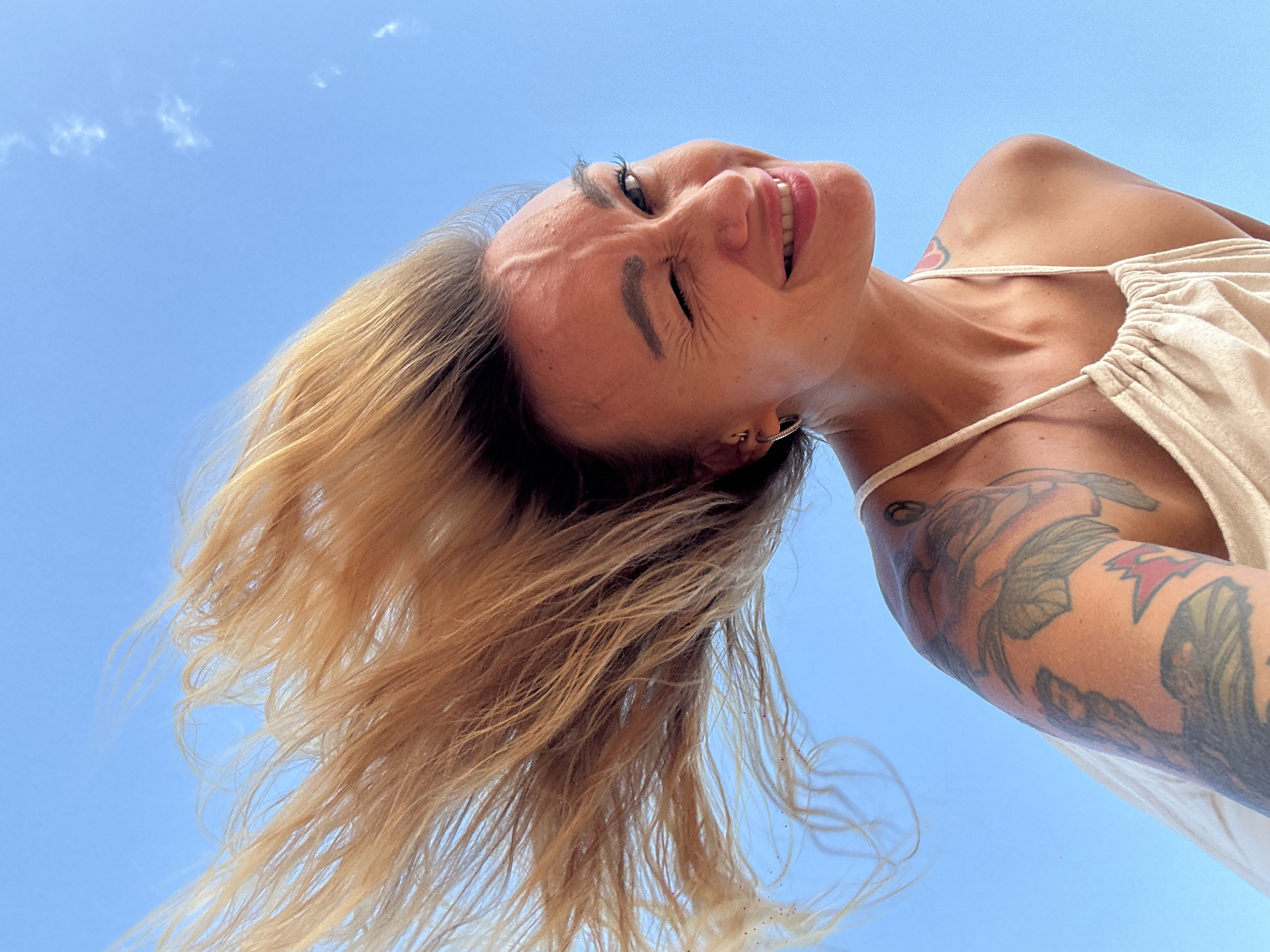 A low-angle shot captures a smiling woman looking upwards against a vibrant blue sky with a few sparse white clouds. Her head is tilted back, revealing a wide, joyful smile and partially closed eyes, suggesting a moment of happiness or laughter. Her long, blonde-to-light brown hair flows around her head, indicating movement or a gentle breeze.
She is wearing a light-colored, possibly beige or off-white, top with thin shoulder straps. Prominent tattoos, featuring floral and possibly other intricate designs, cover a significant portion of her left arm and shoulder, with another small tattoo visible near her right collarbone. A subtle silver hoop earring is visible in her left ear. The bright lighting indicates a sunny daytime setting, likely in Mũi Né, Vietnam. No text is visible in the image.