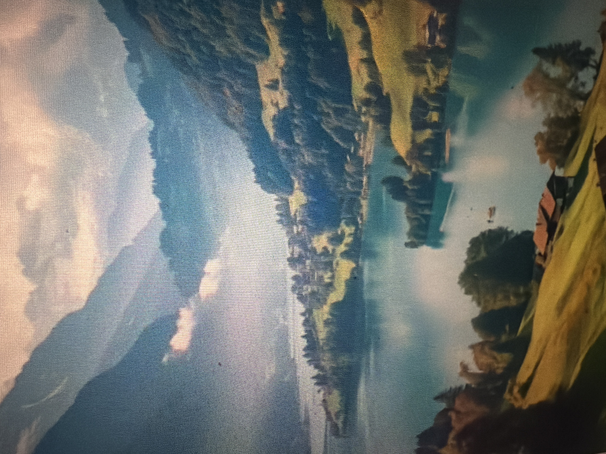 The image depicts a scenic landscape with mountains, a lake, and a village. The mountains in the background are covered in lush green trees, with some patches of light illuminating the slopes. The lake in the foreground is calm and reflects the sky and surrounding scenery. A small village with houses and boats is visible along the shore of the lake. The sky is partly cloudy with soft sunlight filtering through. The overall scene conveys a sense of tranquility and natural beauty. There are no people or specific actions visible in the image. The image is a photograph of a natural landscape, likely a lake and mountain region. The question about Baan Mae Nam, Thailand, is not supported by the visual content of the image.