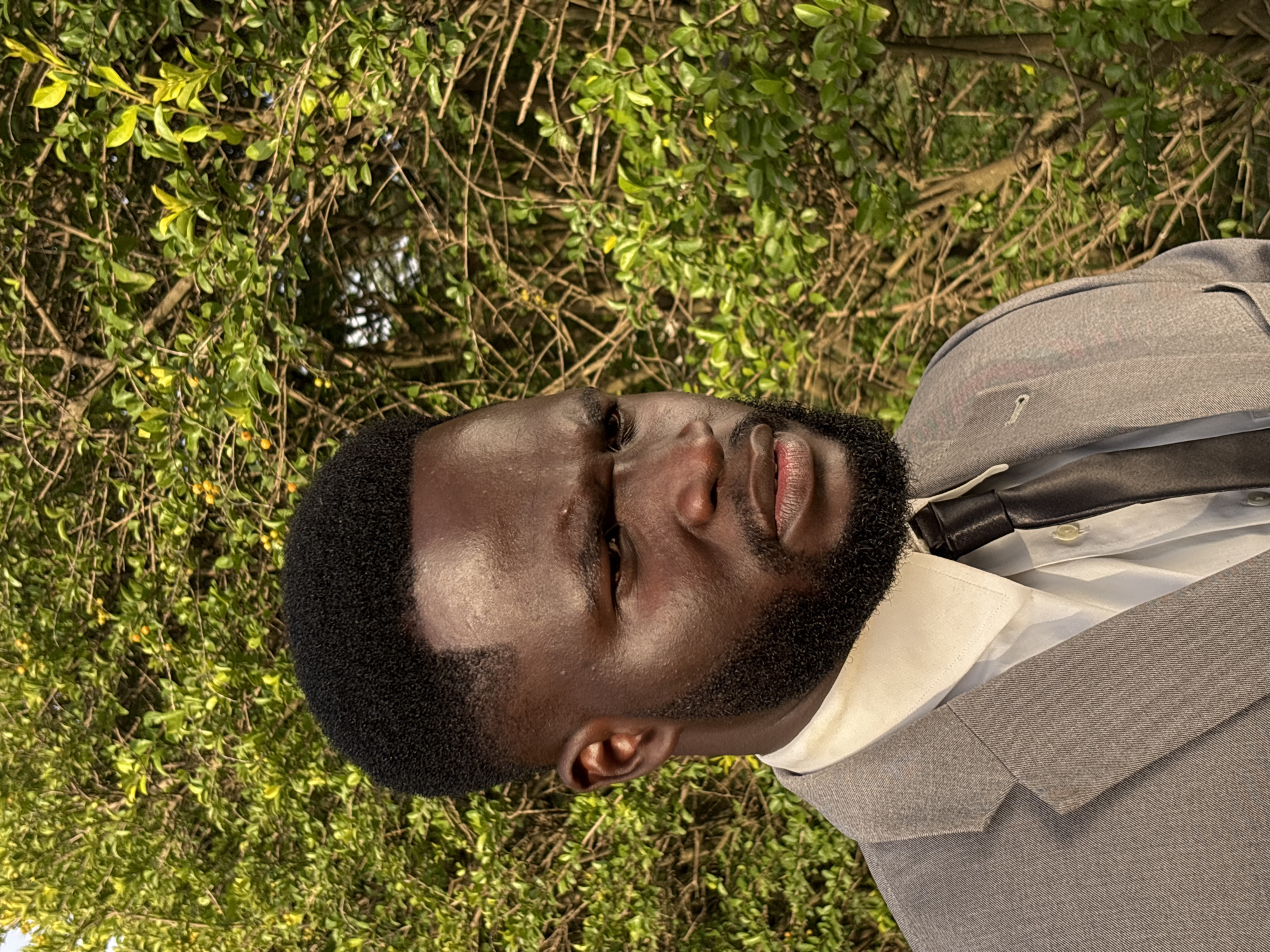 The image is a close-up, portrait-style photograph of a young Black man wearing a gray suit, white dress shirt, and a black tie. His head is tilted back and to the side, with his eyes closed and his lips slightly parted. He has a neatly trimmed beard and short, dark hair. The man is outdoors, with a background of lush green foliage and brown branches, suggesting a natural setting. The lighting indicates it might be daytime, possibly late afternoon given the soft, diffused light. There is no visible text in the image. The overall mood is serene and contemplative. The location context is Bukuru, Nigeria.