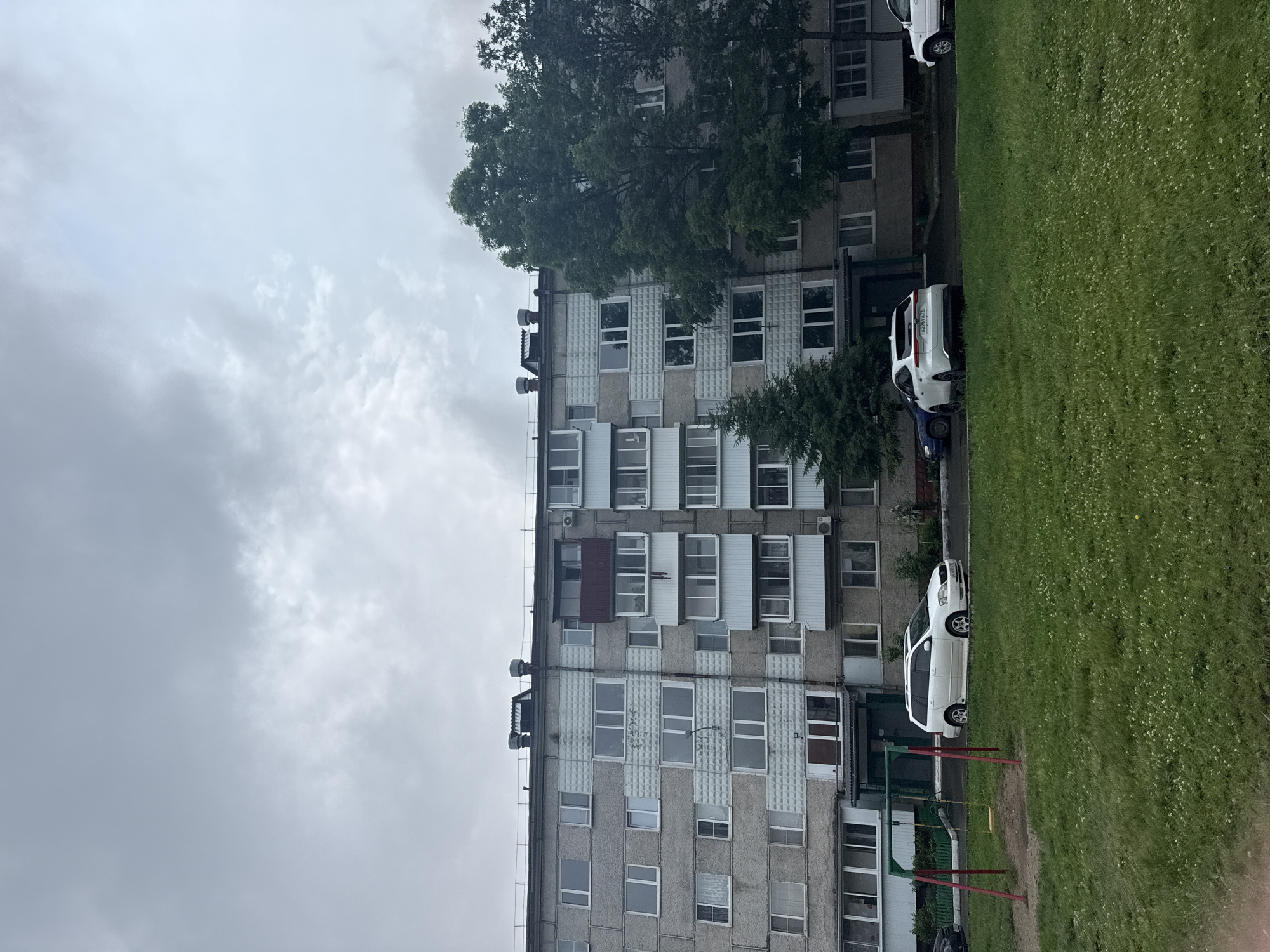 The image captures a residential building in Birobidzhan, Russia, under an overcast sky. The building is a multi-story structure constructed with gray concrete blocks and features numerous windows and balconies. Some balconies have glass enclosures, while others are open. Decorative textured panels are visible on some sections of the building's facade.
In front of the building, there is a grassy area with a children's playground consisting of a swing set. Several cars are parked on a paved area adjacent to the lawn. These include a white station wagon, a white SUV, and a dark blue sedan. Trees, including a large deciduous tree and a coniferous tree, are located near the building and on the right side of the frame. The overall scene depicts a typical urban residential environment.
