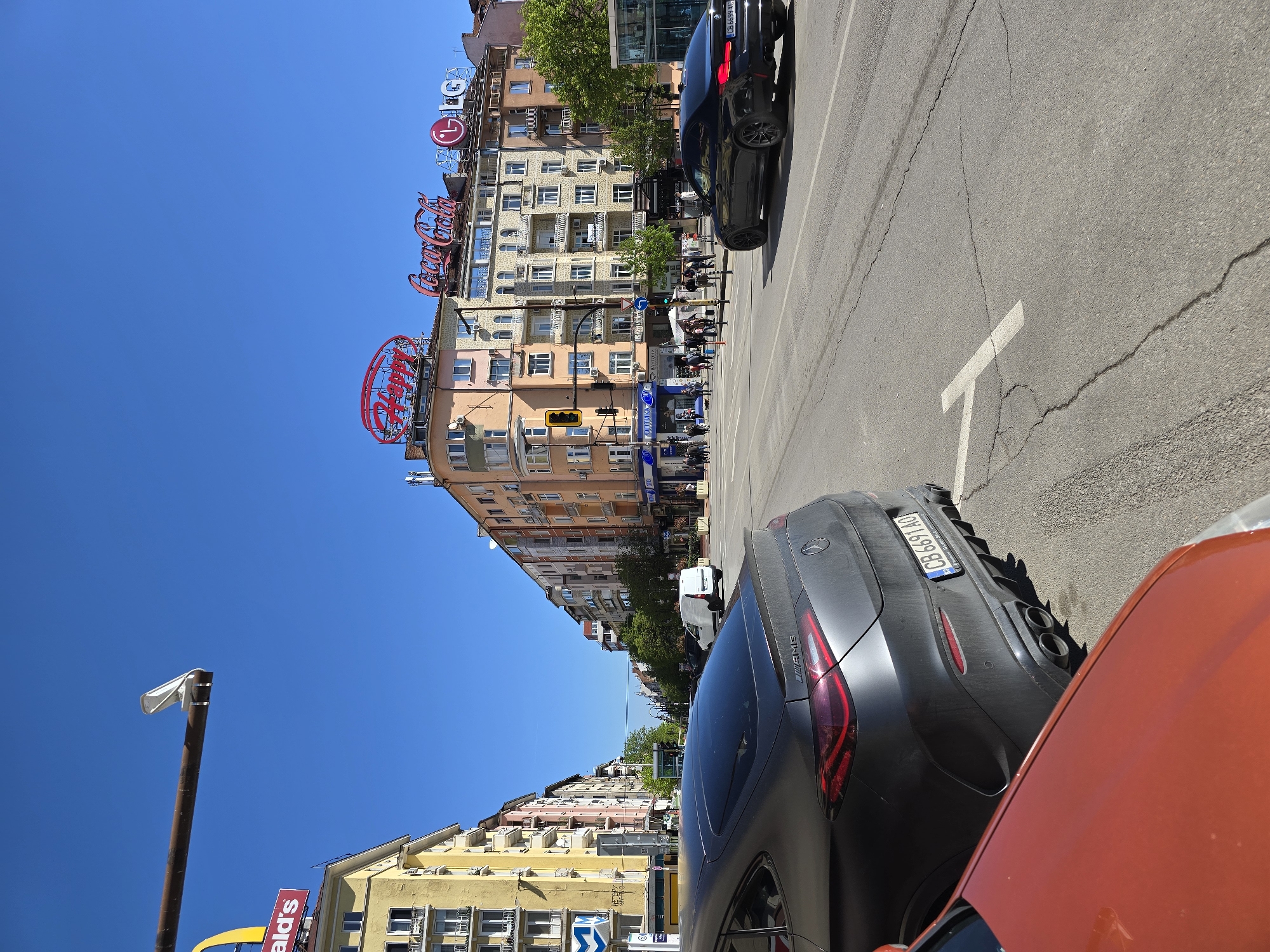 The image is taken on a street in Sofia, Bulgaria, under a clear blue sky. Several buildings line the street, with prominent signage for Coca-Cola, LG, and other brands visible on their facades. A traffic light is mounted to one of the buildings.
The lower part of the image features portions of two cars; one is orange, and the other is a gray AMG model with a license plate number CB 6619 HO. A black car is seen in the distance on the road. Pedestrians are walking on the sidewalks along the street. A street lamp is located on the side of the road.