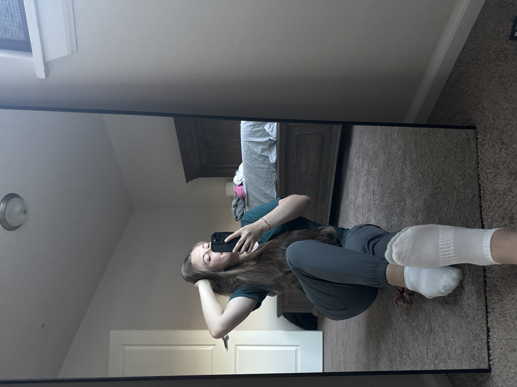 A young Caucasian woman with long, light brown hair is sitting on a carpeted floor, looking into a large floor mirror. She is wearing a dark green scrub top and dark grey jogger-style pants, and white ankle socks. Her left hand is behind her head, and her right hand is holding a black smartphone, which she is using to take a selfie. In the mirror's reflection, a bed with a grey comforter and a dark wooden headboard is visible in the background, along with a doorway to what appears to be a closet. The room is modestly furnished with neutral-colored walls. The overall impression is a casual, domestic scene, likely within a residential setting.