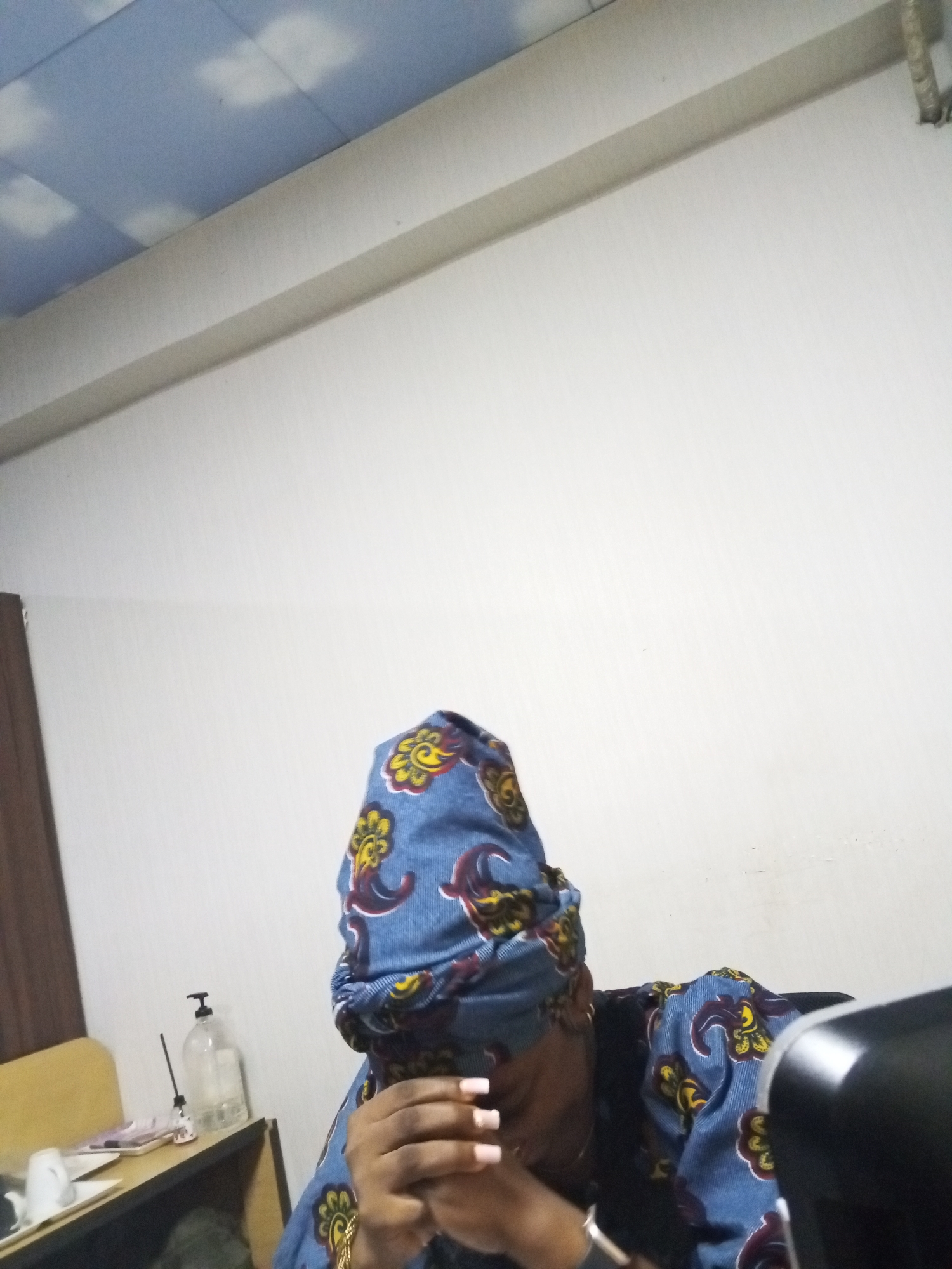 The image depicts a person wearing a blue and yellow patterned headwrap and shirt, with hands clasped together in what appears to be a gesture of concern or contemplation. The person is indoors, situated near a desk cluttered with items including a pump bottle, a white mug, and miscellaneous objects. The background features a plain white wall and a ceiling with cloud-like patterns. There is insufficient information to determine the specific location in Abuja, Nigeria.