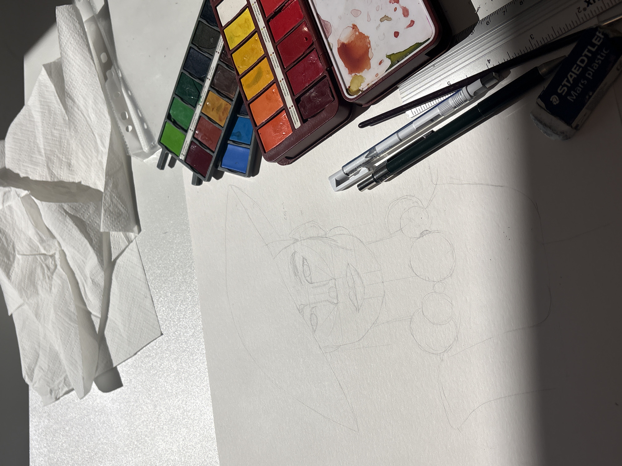 The image depicts an artist's workspace. A sheet of white paper sits partially in shadow. A faint pencil sketch of a cartoon character with large eyes, circular facial features, and a pointed chin is visible on the paper. To the left, there is a crumpled pile of white paper towels. At the top, an open watercolor palette with various colors (green, brown, yellow, orange, red, and blue) is positioned alongside a metal ruler, two pens (one silver and one black), a watercolor brush, and a Staedtler Mars plastic eraser. The location is unknown and may or may not be in the city of Milan.