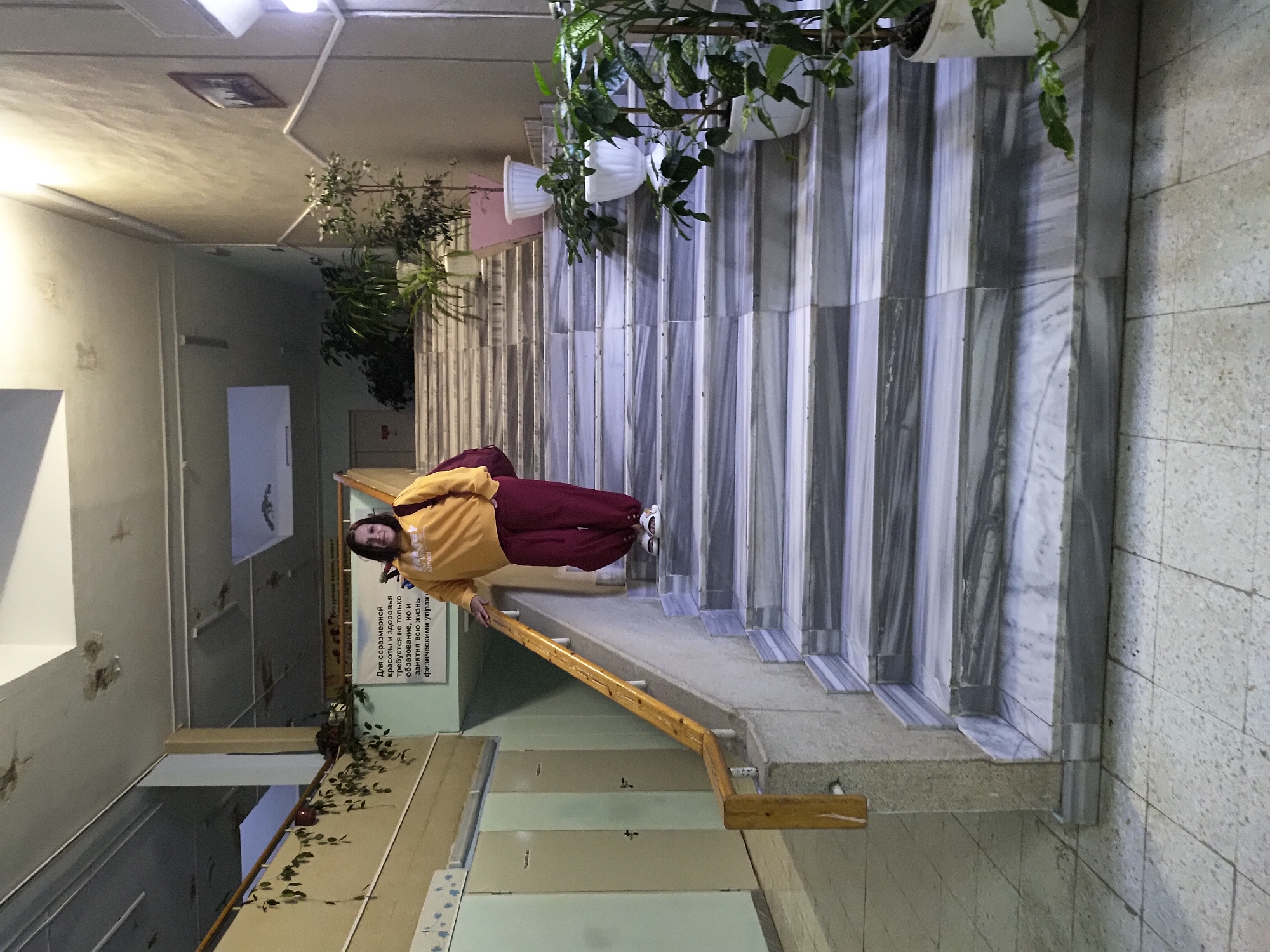 Here is a detailed description of the image:
**Scene:** The image is taken in what appears to be an indoor stairwell or hallway. The dominant features are a set of marble or stone steps, and a handrail along the side. There are plants placed along the top of the steps, possibly in pots.
**People:** A person is positioned on the steps. They appear to be wearing a yellow top/jacket and maroon or reddish-brown pants.
**Objects:**
* Marble or stone staircase.
* Wooden handrail.
* Potted plants with green foliage.
* Signage with text.
**Actions:** The person is standing on the steps, apparently posing for the photo.
**Environment:** The area seems to be within a public building or institution. The walls are light-colored and there are some decorative elements like vines or additional plants near the bottom of the staircase. The lighting seems to be primarily artificial, based on the fixtures visible on the ceiling.
**Location:** The image has a sign which says "Для соразмерной красото и здоровья требуется не только образование, но и занятия всю жизнь физическими упражн. Attract" This indicates that the image is likely taken in Russia or a Russian-speaking area in the city of Unknown, Unknown.