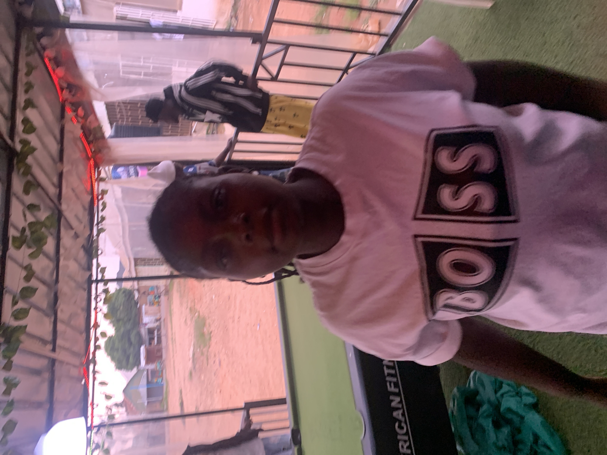 A person wearing a light-colored t-shirt with the word "BOSS" printed on it is the primary subject of this photograph. The scene appears to be on a rooftop or balcony with a pergola-like structure overhead, decorated with artificial foliage and red lights. Another individual is visible in the background wearing a striped shirt. In the foreground, part of a workout machine with the words "American Fitn" is visible. The setting gives a sense of being in an urban residential area, although precise architectural details for definitive location cannot be made. The picture was taken in Jos, Nigeria.