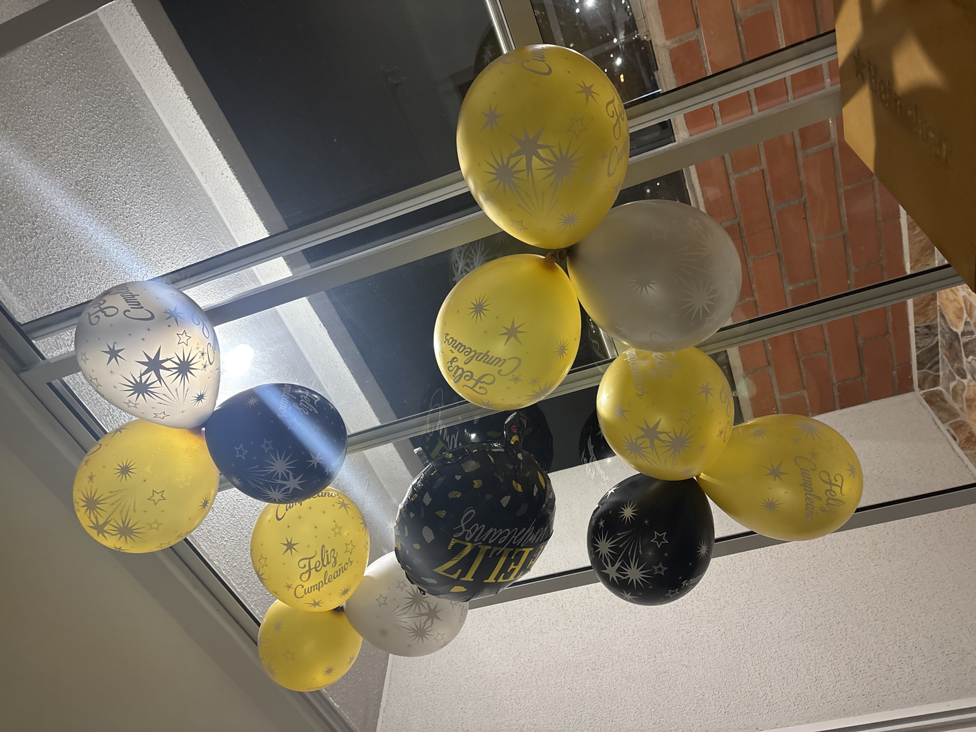 The image depicts an indoor scene in Guarne, Colombia, dominated by a festive arrangement of approximately fifteen balloons. The balloons are primarily yellow, black, and white/silver. Several yellow latex balloons feature silver print displaying the text "Feliz Cumpleaños" alongside starburst and individual star designs. Black latex balloons and white/silver latex balloons are also present, bearing similar silver starburst and star patterns. A single, larger, irregularly shaped black foil balloon prominently displays "FELIZ Cumpleaños" in gold-yellow lettering, accompanied by gold speckles. The balloons are clustered together, appearing to float or be suspended, and occupy a significant portion of the central and upper frame.
The background features a large window or glass door extending vertically, through which a dark exterior, likely a night scene, is visible. Reflections of the balloons and indoor lighting are present on the glass surface. Through the lower section of the window, a reddish-brown brick wall with horizontal mortar lines is discernible. To the upper left, a light-colored wall or ceiling with a subtle textured finish is present. The lower left area of the image shows a section of a tiled floor with light-colored, patterned tiles. In the bottom right corner, a dark, flat surface, potentially a table or box, is partially visible. No individuals are depicted in the scene. The ambient light suggests an indoor setting illuminated by artificial light sources.
