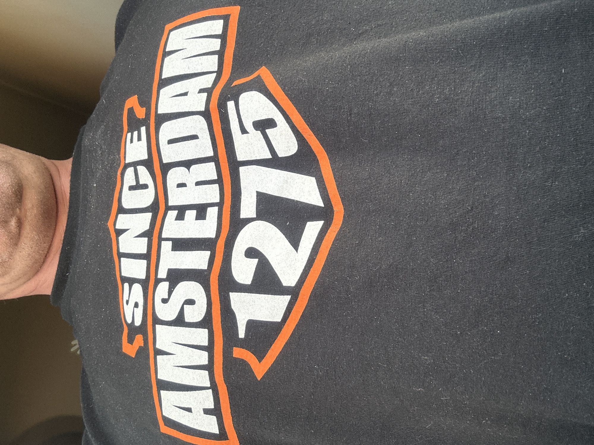 The image shows a close-up of a black t-shirt with a design in white and orange. The design features the words "SINCE AMSTERDAM" stacked vertically, with the number "1275" to the right. The design is enclosed within an orange outline. The photo includes part of a person's neck and skin. The image does not show Sofia, Bulgaria.