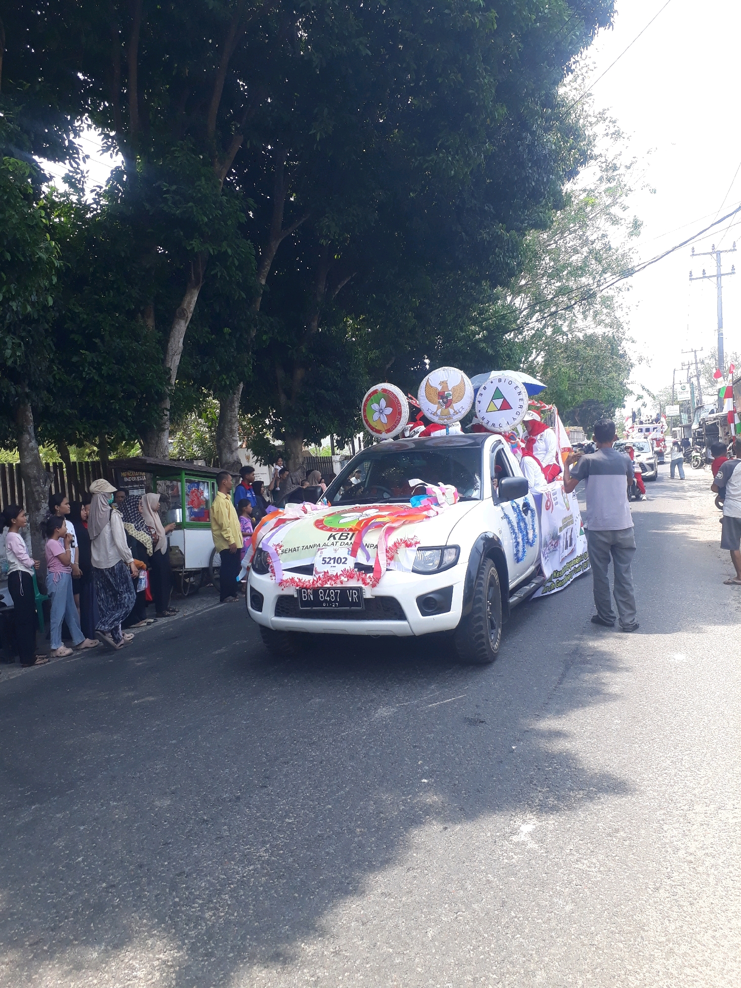 A white pickup truck decorated for a parade is driving down a paved road lined with trees. The truck has balloons on top and colorful streamers attached to the front. Several people are standing on the side of the road, watching the parade. A man in a gray shirt and pants is standing on the right side of the road, holding a camera and appearing to film the truck. Another man in a gray shirt and black pants is standing behind the truck. A food cart is visible on the left side of the road. The environment appears to be a sunny day in a city in Indonesia, likely Pangkalpinang.