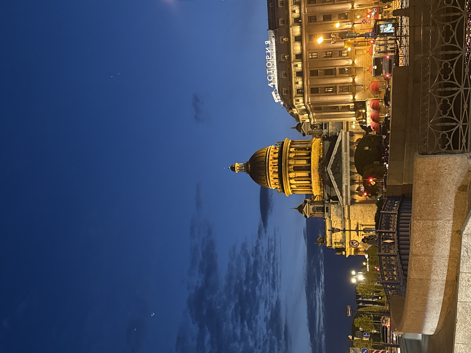 The image displays an illuminated cityscape at dusk or early night in Saint Petersburg, Russia. The sky is a deep twilight blue with scattered, soft clouds.
On the left side of the frame, the prominent golden dome and classical facade of St. Isaac's Cathedral are brightly lit, standing out against the darkening sky. To the right, across what appears to be a large square or thoroughfare, another grand, multi-story building is visible. This building is also well-lit, with numerous windows glowing. On its upper facade, an illuminated sign in Cyrillic clearly spells "АСТОРИЯ" (ASTORIA), identifying it as the famous Astoria Hotel. Below the hotel's facade, several distinctive red awnings are visible, likely covering outdoor seating for a restaurant or cafe.
In the immediate foreground on the right, a sturdy stone balustrade or parapet with intricate black metalwork forms a barrier, possibly part of a bridge or an embankment. At street level, beneath the prominent buildings, elements of urban life are present, including a red traffic light, other streetlights, and the partial view of vehicles such as a bus and cars. The overall scene evokes a serene and majestic evening atmosphere in a historic city, with architectural landmarks beautifully highlighted by artificial light.