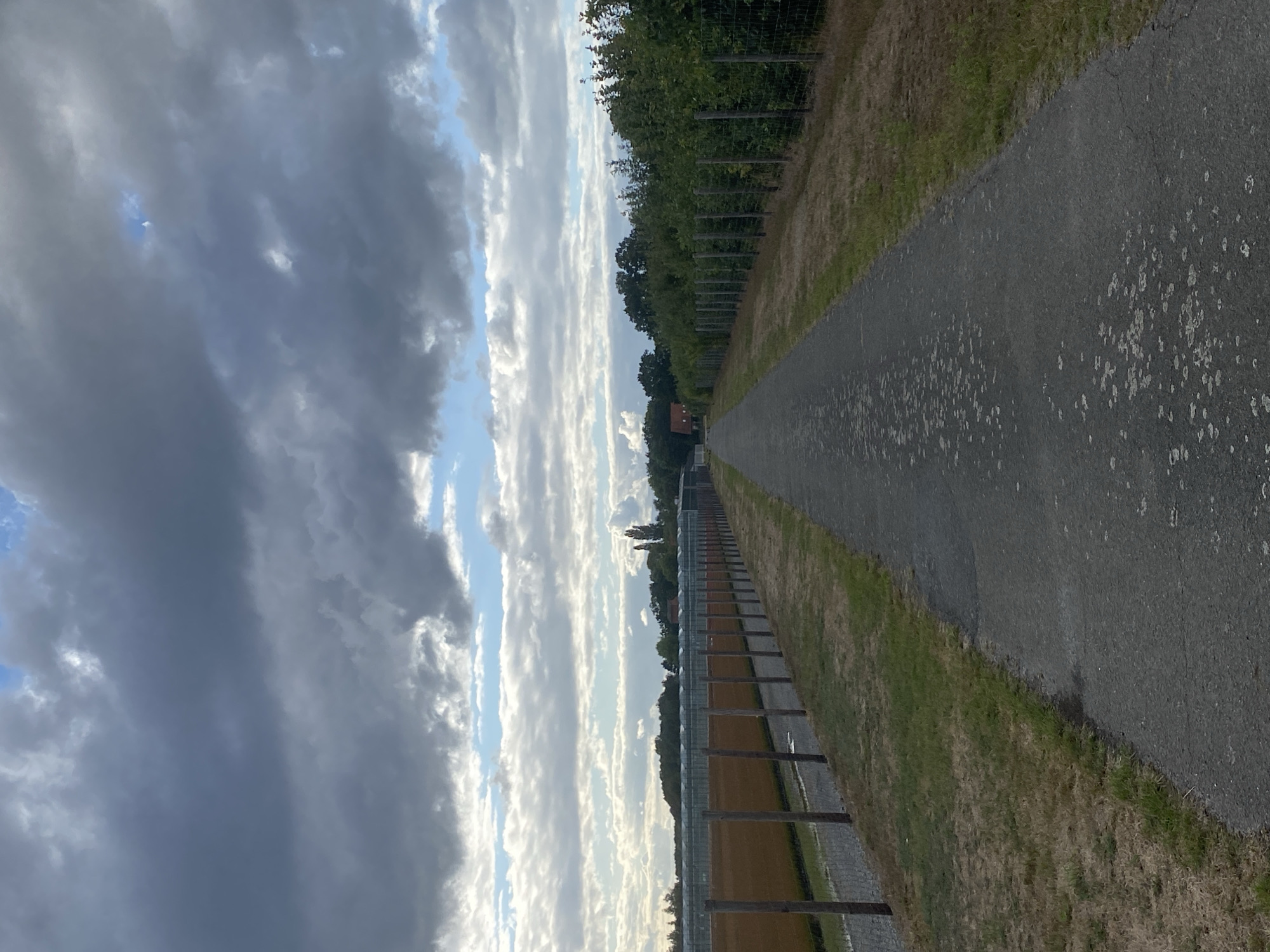 The image depicts a straight, paved pathway leading into the distance, bordered by a fence and dry grass on the right, and a fence, gravel path, and a field with wooden posts on the left. In the background, a line of trees and what appear to be greenhouses or agricultural structures are visible. A small, red-roofed building is situated among the trees. The sky above is filled with dramatic, dark clouds with patches of bright blue sky peeking through. The overall scene suggests a rural or semi-rural environment, possibly an agricultural area. The location is identified as Schwarme, Germany.