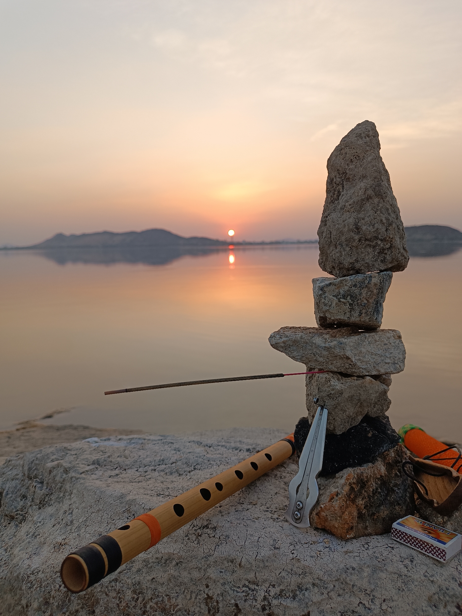 The image depicts a serene scene at a body of water, likely a lake or river, near Kammavaripalli, India, as suggested by the camera location. The sun is setting, casting a warm glow over the water and distant hills. In the foreground, a stack of rocks is arranged in a cairn. An incense stick is placed on the stack, seemingly burned. Nearby, a wooden flute lies on a light-colored rock surface. A jaw harp is positioned near the base of the rock stack, along with a small percussion instrument with an orange cover and a box of "SUNVILI" brand wax matches. The scene is peaceful and suggestive of meditation or a moment of reflection. There are no people visible. The composition emphasizes the harmony between nature and handcrafted objects.