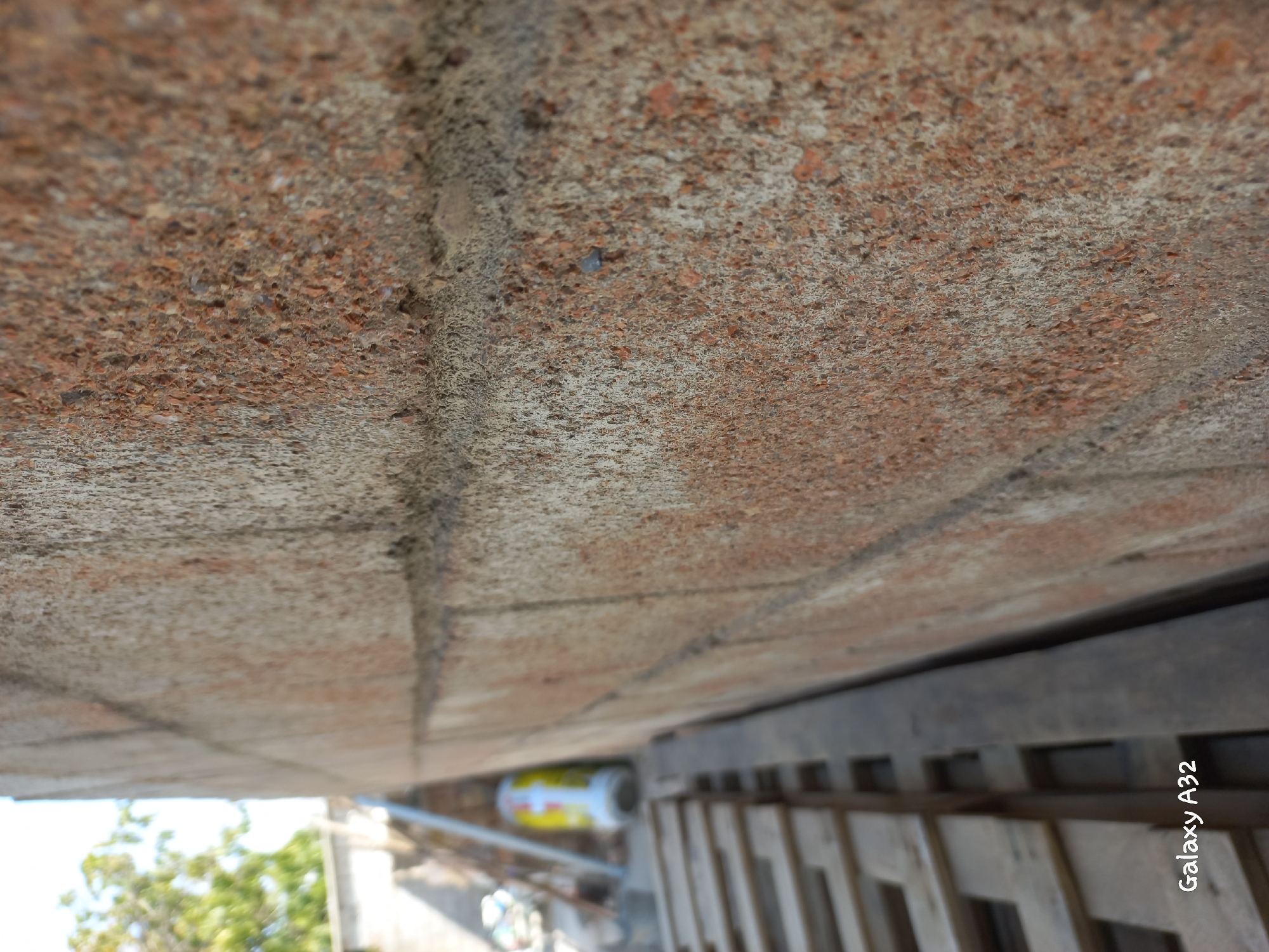 The image displays a close-up view of a textured brick wall, with a shallow depth of field. The bricks are a reddish-brown color with a coarse, gritty surface. The mortar between the bricks is visible and appears to be lighter in color. In the background, a blurred scene suggests an outdoor environment, possibly a construction or storage area. To the left, a wooden pallet structure is partially visible, and further back, a yellow and white cylindrical object, possibly a container or tank, can be discerned. Some greenery and a hint of sky are present in the far distance, out of focus. The watermark "Galaxy A32" is in the lower left corner. The scene does not provide any specific indicators of the location being Kemin, Kyrgyzstan.