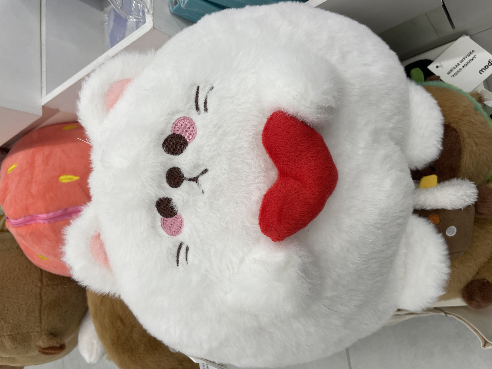 The image shows a close-up of several plush toys arranged on a shelf. The main subject is a white, round plush toy resembling a cat or bear, with embroidered facial features including closed eyes, pink cheeks, and a red heart-shaped tongue sticking out. To the left, partially visible, is a round orange plush toy with yellow spots, resembling a fruit or dessert. To the right, and behind the white plush, are other plush toys, one of which appears to be brown and green, possibly representing a food item like a roll or sushi. A tag with Cyrillic text and the brand name "modi" is visible on the right side of the image. The toys are soft and fuzzy, suggesting they are intended for children or as decorative items. The setting appears to be a retail environment, likely a store, given the presence of shelves and product packaging in the background. The lighting is bright, indicating it is daytime. The overall mood is cheerful and playful, conveyed by the cute design of the plush toys. The Cyrillic text on the tag reads "МЯГКАЯ ИГРУШКА "РОЛЛ-РОЛЛИН"", which translates to "SOFT TOY 'ROLL-ROLLIN'".