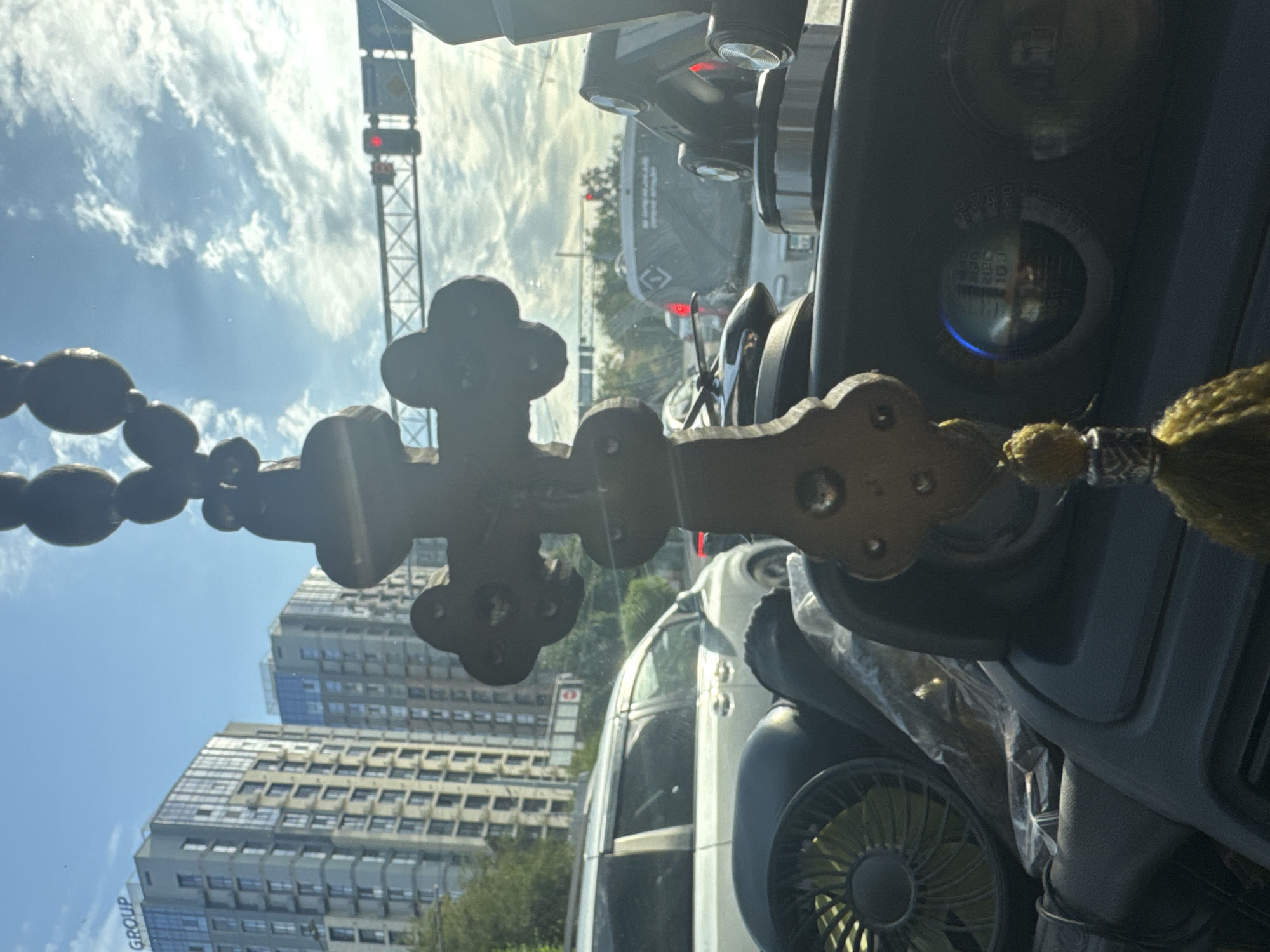 The image, taken from inside a vehicle in Almaty, Kazakhstan, showcases a close-up view of a beaded religious cross hanging from the rearview mirror. The cross, made of dark wood or beads, has a decorative element at its center. In the background, through the windshield, a bustling cityscape is visible. Two tall, modern apartment buildings with reflective glass facades dominate the left side of the frame, with the word "GROUP" clearly visible on the rooftop of the building on the far left. The sky above is a bright blue, dotted with fluffy white clouds. Traffic lights and other road infrastructure are also visible, indicating the vehicle is in motion or stopped at an intersection. Several cars are on the road, including a white minivan to the left and a dark SUV with text on its rear window further down the road. The interior of the car features a dashboard with circular gauges, one of which is illuminated blue. A small, circular fan is also visible on the left side of the dashboard, partially obscured by a plastic bag. The overall scene captures a typical urban environment in Almaty under a partly cloudy sky.