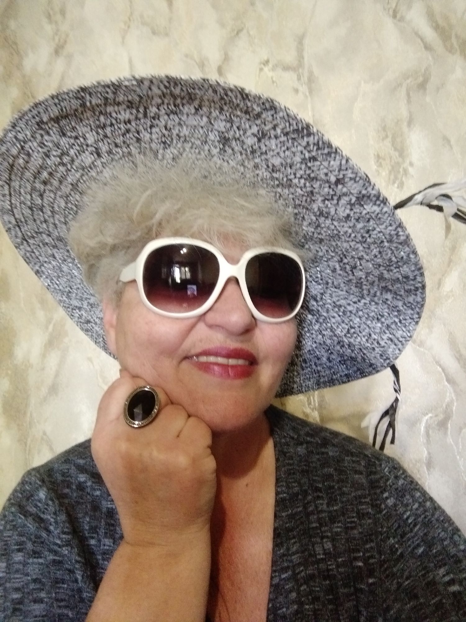A mature woman wearing an oversized speckled hat and large white-framed sunglasses is posing for a picture. She rests her fist, adorned with a large black ring, against her chin. She wears a heathered gray cardigan. The background is a textured wall. The location is Horishni Plavni, Ukraine.