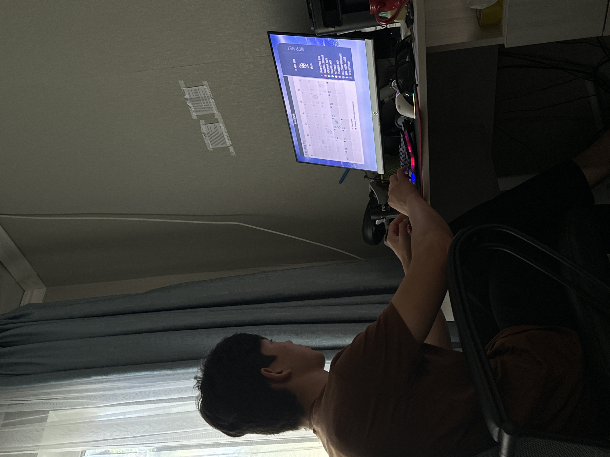 A young male is seated at a desk, facing a computer monitor displaying a calendar and a list of football matches. The man is wearing a brown t-shirt and black pants. He is actively using a keyboard. The desk is cluttered with various items, including a mouse, headphones, and a red mug. Behind the desk, blue curtains are drawn across a window, and the wall features two pieces of paper taped up. The scene appears to be a home or office environment, possibly in the city of Temirtau, Kazakhstan, given the context provided. The lighting suggests it is daytime, with natural light filtering through the curtains.