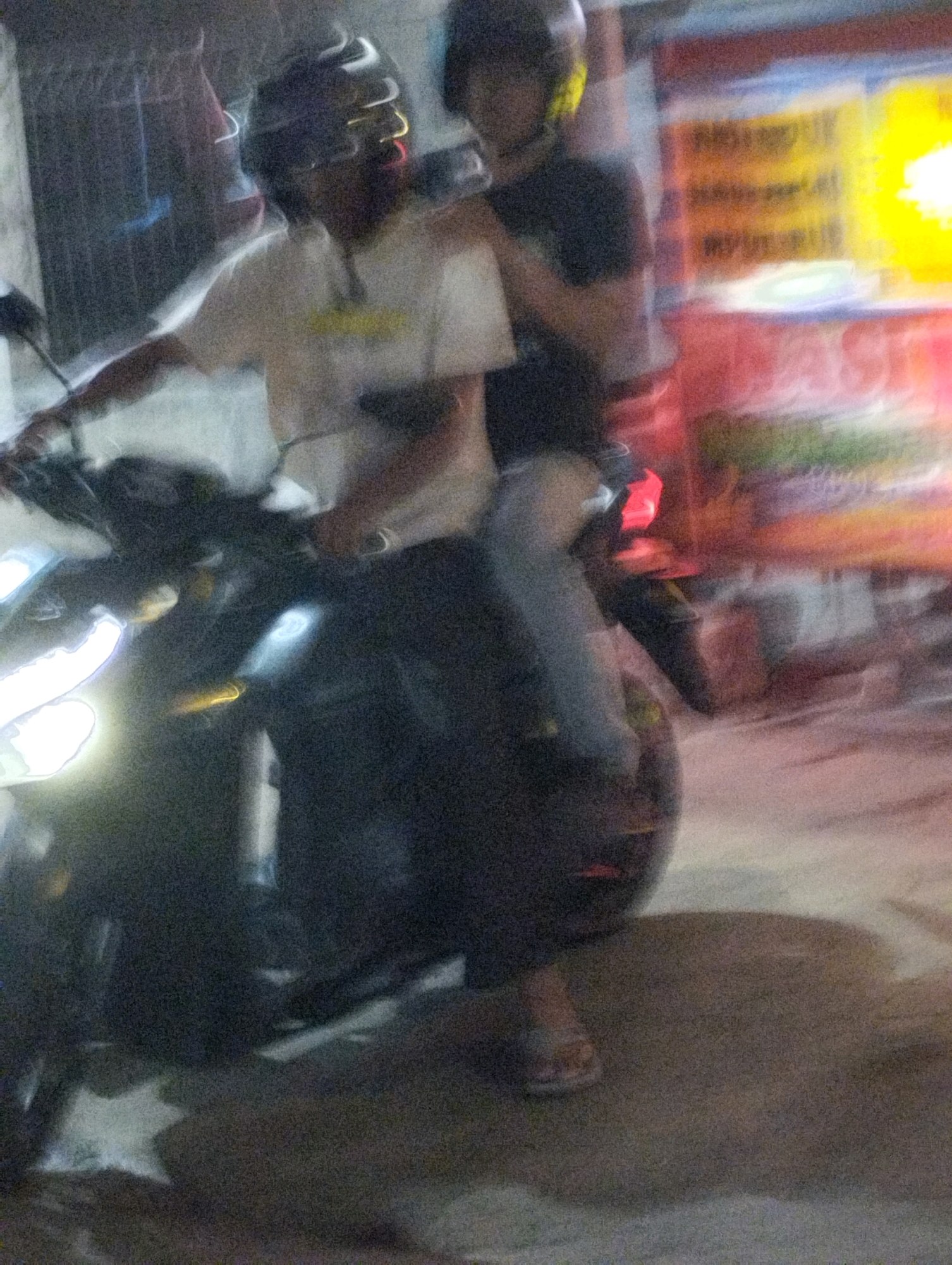 In Bekasi, Indonesia, two individuals are captured in motion on a motorcycle. The person in front, wearing a helmet with reflective white and yellow accents, is dressed in a light-colored t-shirt and dark pants. They are holding the handlebars of the motorcycle. Behind them, another individual also wearing a helmet, this one dark with yellow accents, is seated. This person is wearing a black t-shirt and dark pants, with their arms resting on the person in front. The motorcycle itself appears dark with illuminated elements, including a bright white headlight and what seems to be a yellow indicator light. The background is blurred, suggesting movement, and includes indistinct shapes of buildings or stalls with glowing signage in vibrant colors, primarily yellow and red, indicative of a nighttime urban setting in Bekasi. The ground beneath the motorcycle is paved and somewhat textured. The overall scene is characterized by motion blur and low light conditions.
