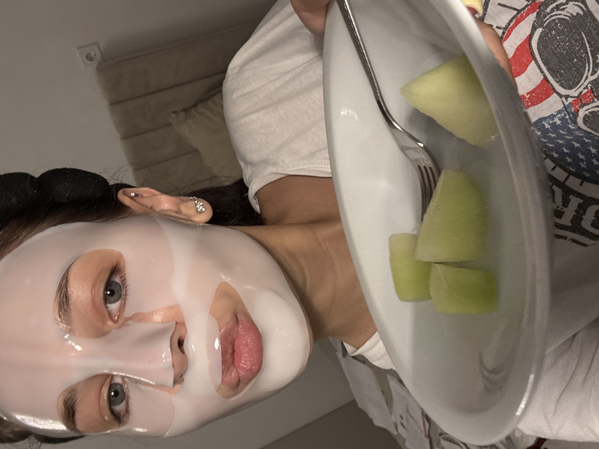 An indoor scene in Rakovski, Bulgaria, depicts a female subject, estimated to be in her twenties or thirties, captured in a chest-up, slightly low-angle photograph. She directly faces the camera with her lips subtly puckered. Her face is covered with a translucent white sheet mask, exposing her eyebrows, blue-grey eyes, and pinkish lips. Her dark hair is partially visible on the right side of her head, styled in a bun or rollers. Her left ear features a helix piercing and a small, sparkling bow-shaped earring in the lobe. She wears a white short-sleeved t-shirt. The subject holds a white, shallow bowl-like plate containing four irregular pieces of light green melon and a silver-colored metal fork. In the background, a beige upholstered headboard and a matching pillow are visible. A white electrical wall outlet is mounted on the light-colored wall above the headboard, positioned on the right side of the frame. A patterned fabric, featuring elements resembling an American flag (red stripes, blue field with white stars) and a black skull graphic, is partially visible in the bottom right corner of the image. The scene is illuminated by soft, ambient light.