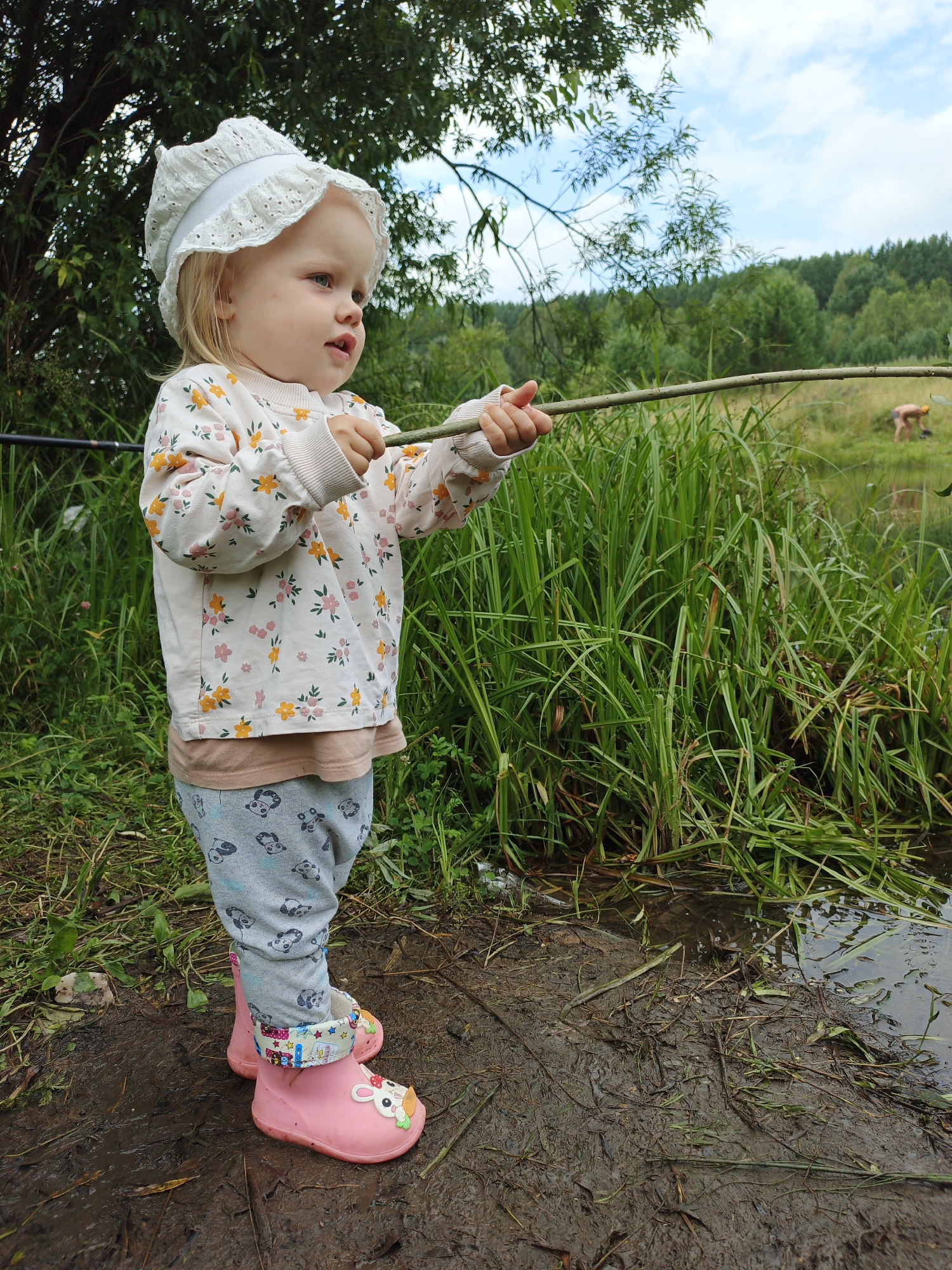 A young Caucasian child, approximately 2-3 years old, stands in muddy terrain next to a body of water. The child is wearing a white lace bonnet, a long-sleeved shirt with a floral pattern of yellow and pink flowers, light grey pants with a panda pattern, and pink rubber boots decorated with animal faces. The child is holding a stick horizontally with both hands, appearing to be playing or fishing. In the background, tall green grass and reeds line the water's edge. Beyond the vegetation, a treeline of deciduous trees forms the horizon under a cloudy sky. Further in the distance, two adult figures are visible in the water, one appearing to be engaged in some activity. The overall environment suggests a rural or natural setting, possibly in Medvedyovka, Russia, given the context of the prompt.