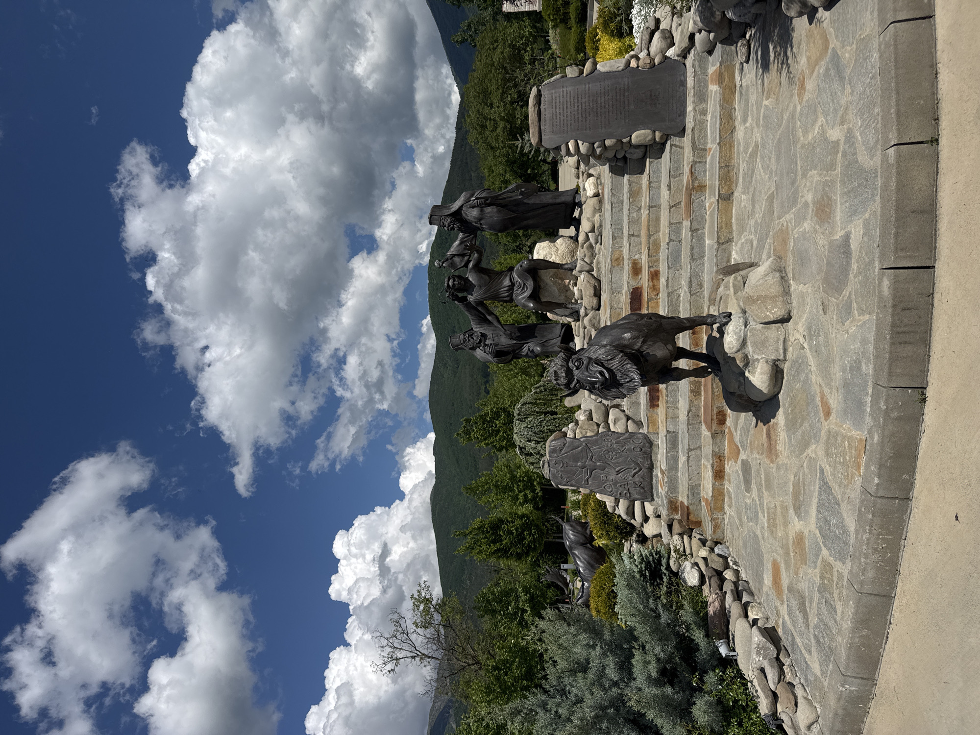 The image shows a monument in Skobelevo, Bulgaria, featuring a bronze sculpture of a group of men and a dog on a stone and rock-lined pathway. The pathway leads up a slight incline, with stone steps visible. The background features green mountains and a blue sky with large, puffy white clouds. The sculpture appears to depict a scene of historical or cultural significance, judging by the dress of the figures and the presence of plaques with inscriptions. Vegetation, including trees and shrubs, surrounds the monument. The scene is outdoors, likely on a sunny day.