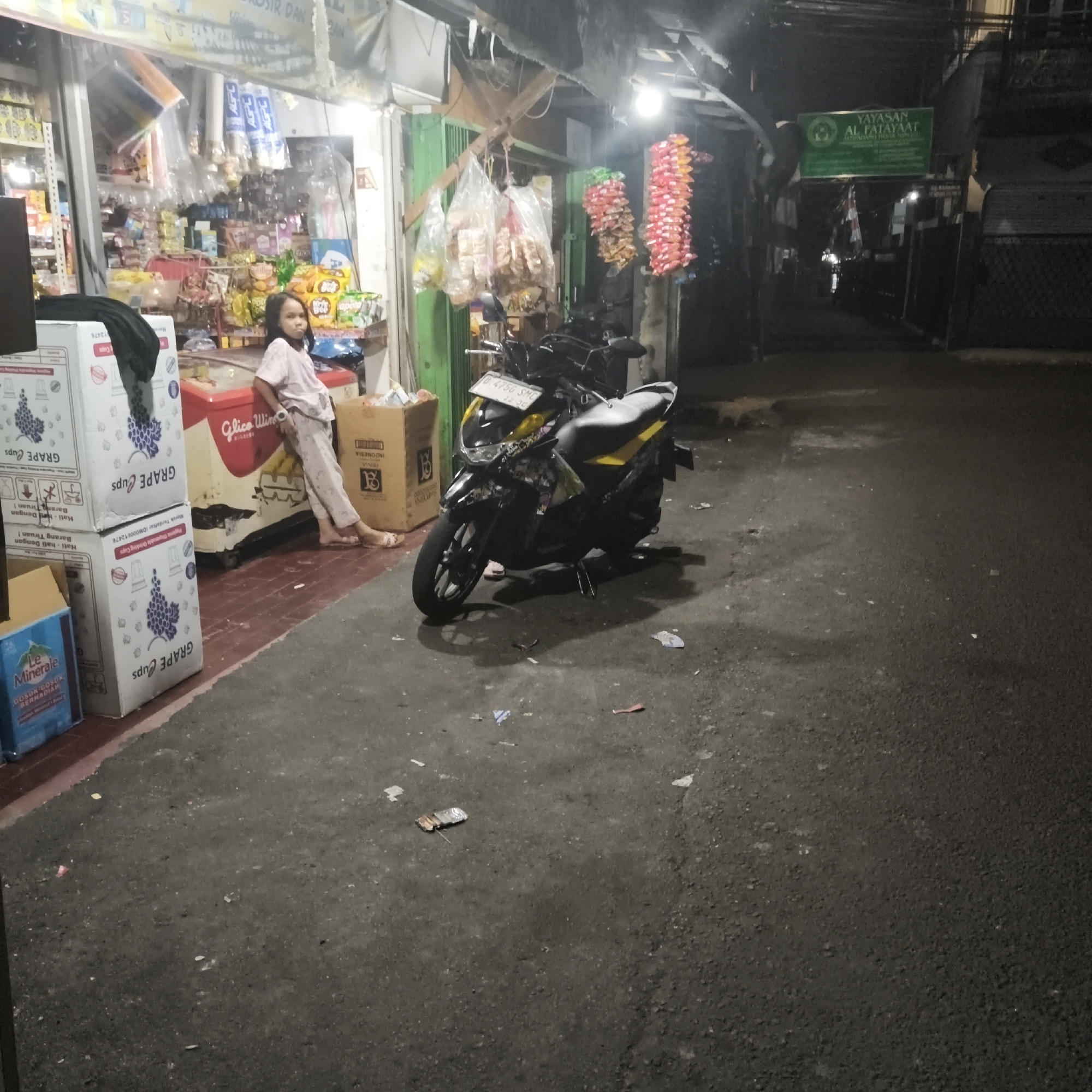 A young girl in a light pink t-shirt and pants leans against a red refrigerator in front of a store stall. The store appears to be selling snacks and drinks, with shelves of products visible behind her. Stacks of cardboard boxes with grape imagery are to her left. A black and yellow motorcycle is parked in front of the stall, with its license plate reading B 475G SMC. Several plastic bags of snacks hang from the awning above the store. In the background, a green sign reads "YAYASAN AL FATAYAA", indicating a foundation. The scene takes place on a paved street, with debris scattered on the ground. The lighting suggests it is nighttime or dusk. This scene is located in South Jakarta, Indonesia.