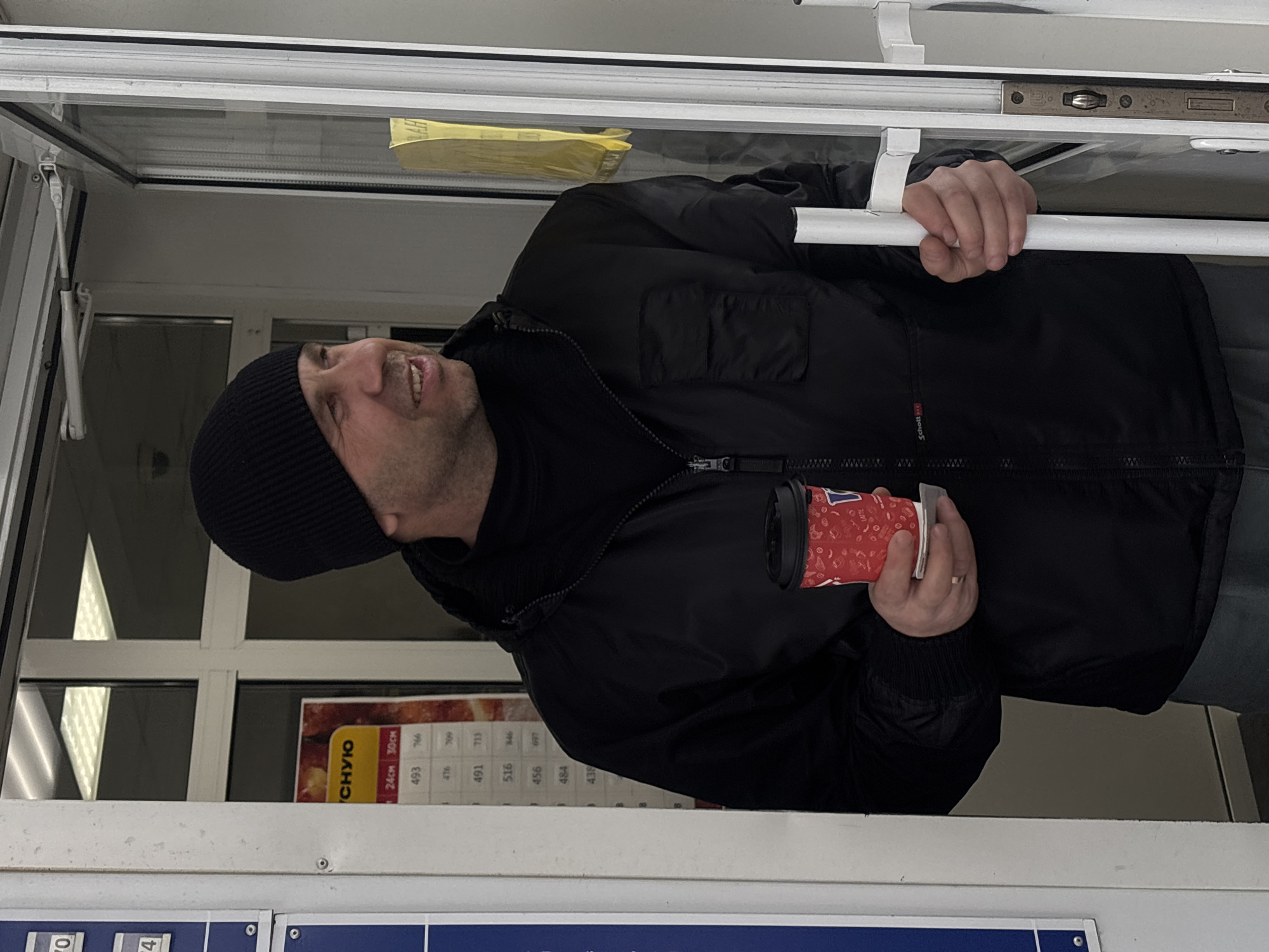 A man is standing at a window, possibly at a service hatch or a small business establishment. He is wearing a black beanie, a black zip-up jacket with a hood, and gray jeans. He is holding a red and white disposable cup, likely a coffee or beverage cup, in his right hand. His left hand is gripping a white metal bar, which appears to be part of the window frame.
The window has a price menu with a word written in Russian "СНУЮ" and prices listed in columns such as 24cm 30cm for the prices 493, 766, 476, 600, 491, 713, 516, 846, 456, 669, 484, 438. The window and surrounding structures appear to be made of white plastic or metal. The location is in the city of Chaikovsky, Russia.