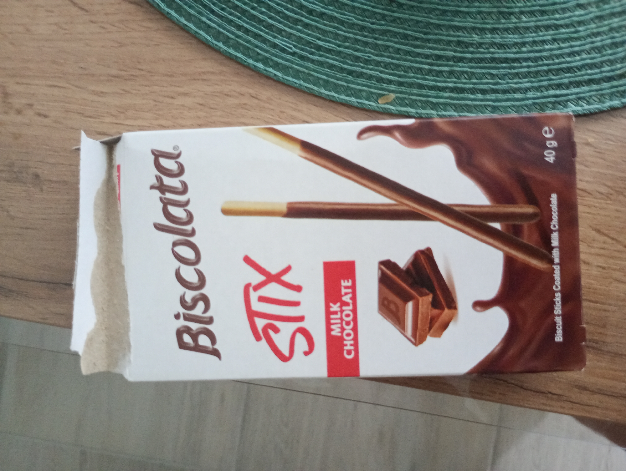 The image shows a package of "Biscopata Stix Milk Chocolate" lying on a wooden surface. The package is partially opened and shows a depiction of chocolate-covered biscuit sticks and pieces of chocolate. The package also has text indicating "40 g e" and "Biscuit Sticks Coated with Milk Chocolate". A green woven placemat is visible in the upper portion of the image. The image does not provide any information about the city of Burgas, Bulgaria.