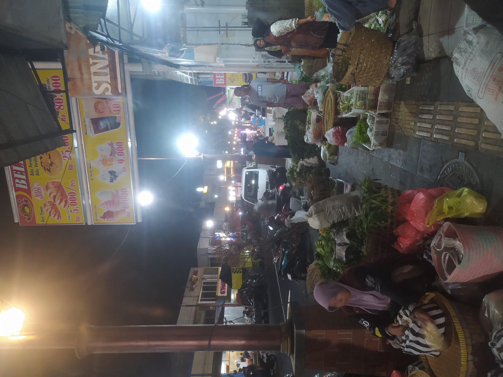 The image depicts a nighttime scene at a street market in Kupang, Indonesia. A prominent street lamp illuminates a portion of the scene, casting a warm glow. In the foreground, a woman wearing a purple hijab and dark jacket is seated, arranging items in a striped bag. She is surrounded by various produce, including leafy greens and what appear to be tomatoes, displayed in woven baskets and plastic crates. Red and yellow plastic bags are also visible.
Behind the woman, the market extends into the background with several stalls and people. A white van is parked in the middle ground, and various motorcycles are scattered around. Overhead, large advertising banners are displayed, one promoting "BELI" with images of bread and another advertising ice cream cones and drinks. The banners also include pricing information in Rupiah.
Further in the background, other market-goers and vendors are visible, engaged in activities typical of a night market. The environment appears to be a paved street with some structures and lighting in the distance. The overall atmosphere suggests a bustling local marketplace operating after dark.