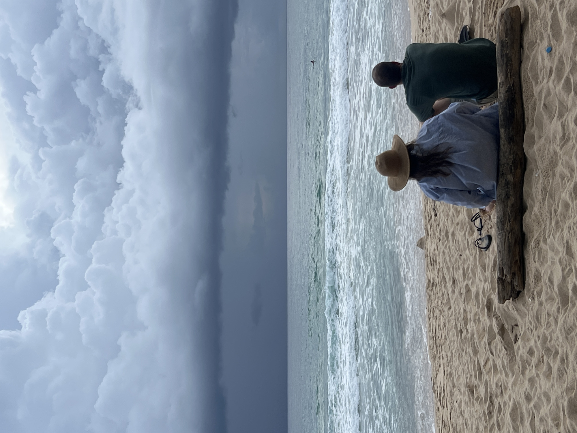 The image shows two people sitting on a sandy beach, facing the ocean. The person in the foreground is wearing a blue and white striped shirt and a straw hat, with their back to the camera. The person behind them is wearing a dark green shirt, also with their back to the camera. They are sitting near a piece of driftwood. The ocean is on their right, with waves breaking on the shore. The sky on the left is filled with dramatic, dark storm clouds, suggesting it might be late afternoon or evening, with a storm approaching. The overall mood is one of calm observation, despite the ominous clouds. The location is identified as Choeng Thale, Thailand. There is no visible text in the image.