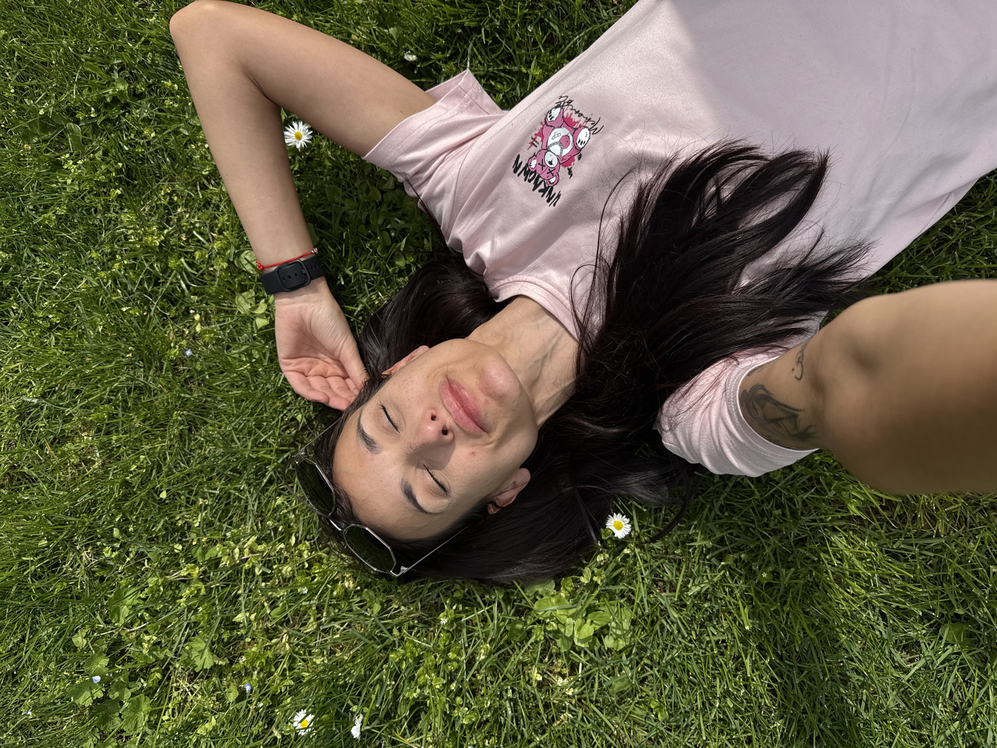 A woman lies on her back in a grassy area. She wears a light pink t-shirt with a cartoon graphic on the upper left side. Her dark hair is spread around her head. She has sunglasses perched on her forehead, and her eyes are closed. She has a black watch on her left wrist and a tattoo on her right forearm. Small white flowers are scattered in the grass around her. The photograph appears to be taken from an overhead perspective, possibly a selfie. There is no way to determine if this image was taken in Pleven, Bulgaria based solely on the visible content of the image.