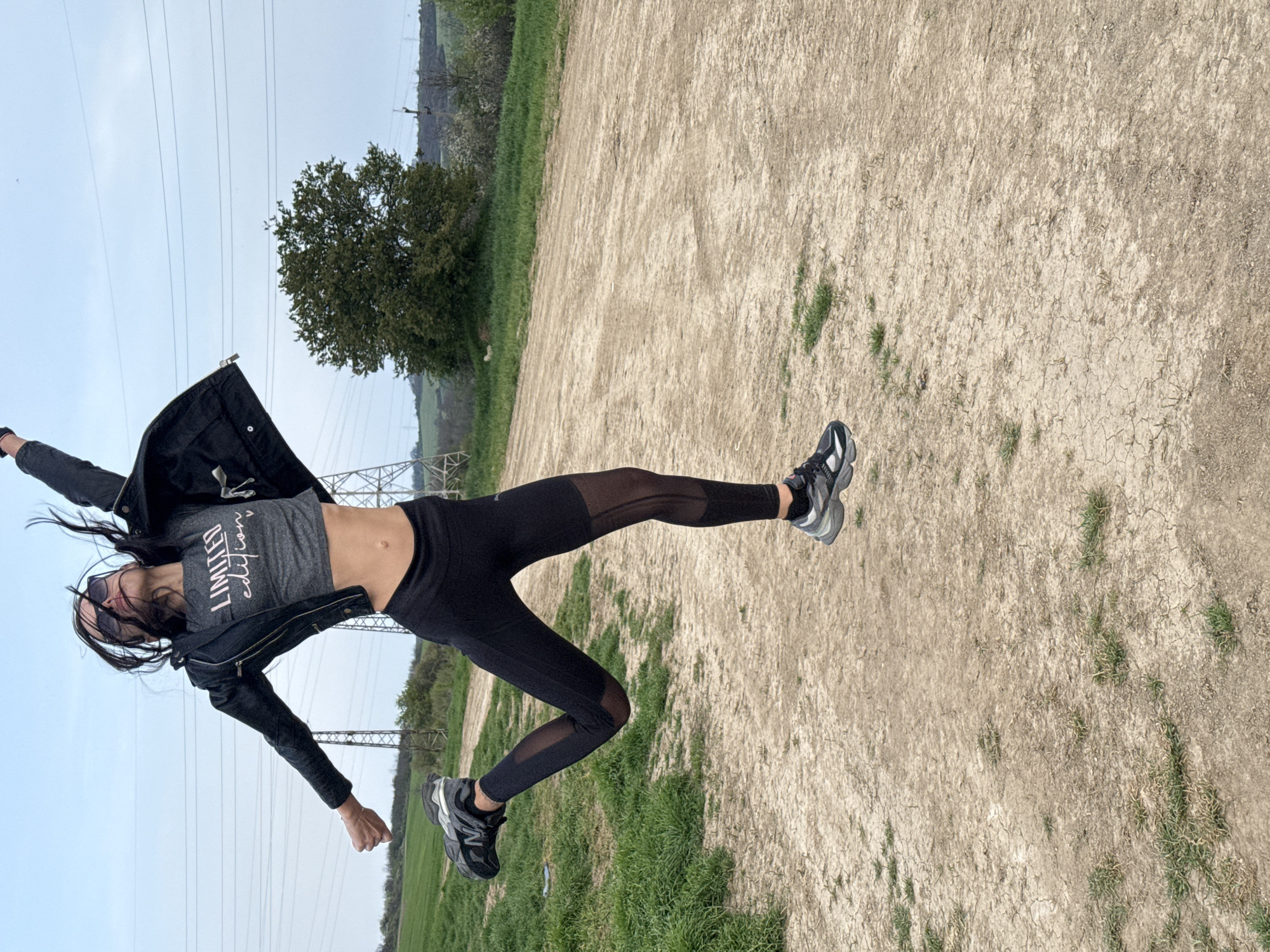 A woman is suspended horizontally in mid-air, appearing to jump or pose dynamically. She wears black leggings with mesh panels, a gray t-shirt with the words "LIMITED edition" on it, a black jacket, and sneakers. Her hair is loose and flowing. She's located in an outdoor environment with a dry, cracked ground surface and patches of green grass. Power lines and pylons are visible in the background, as well as trees and a distant hill. The sky is overcast. There's no clear indication of the city of Pleven, Bulgaria in this image.