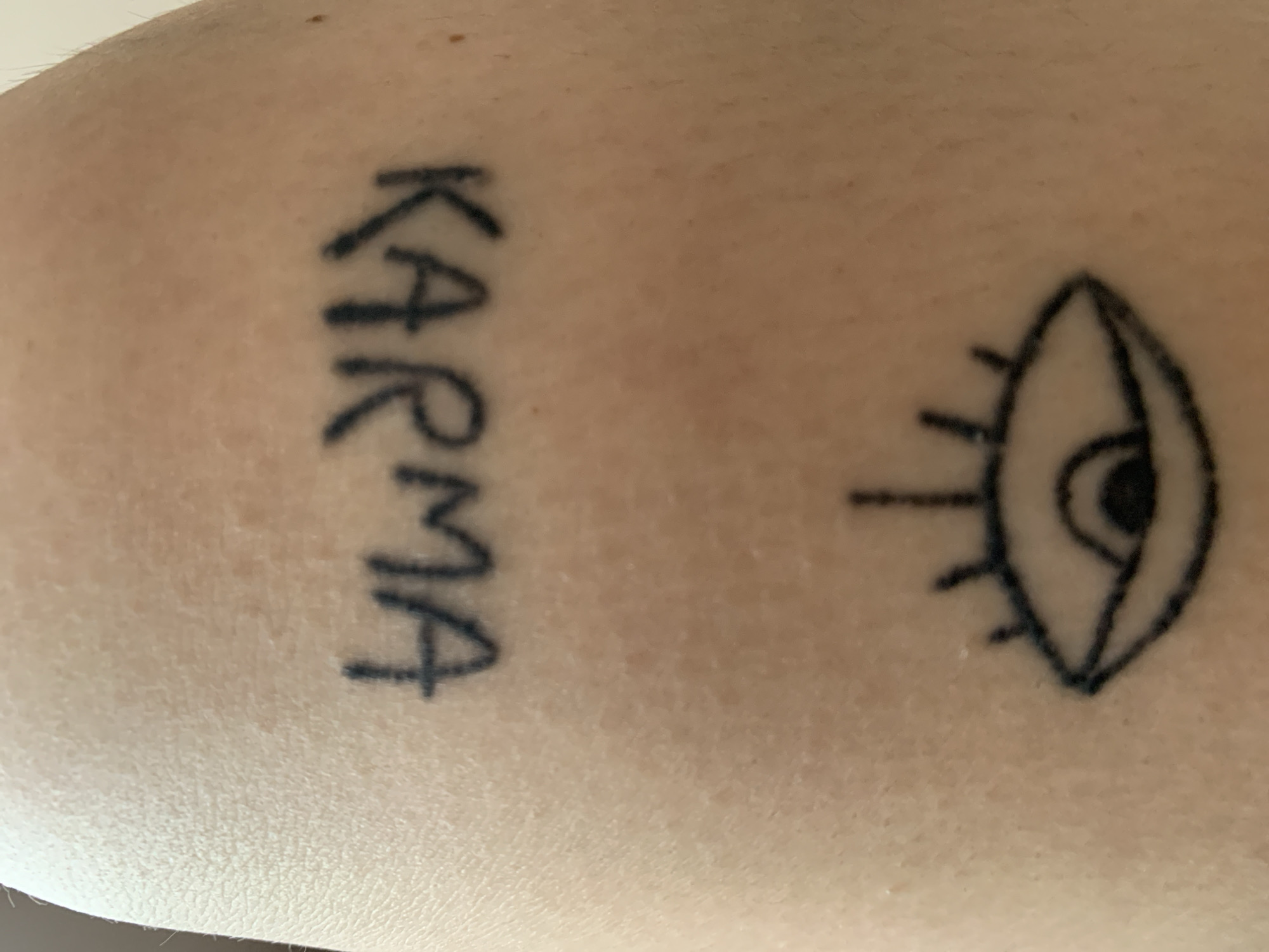 A close-up photograph shows a person's light-skinned arm with two tattoos. On the left side is the word "KARMA" tattooed vertically in capital letters. To the right of the word, there is a simple line-drawing tattoo of an eye with short lines representing eyelashes. The background is the skin of the arm.