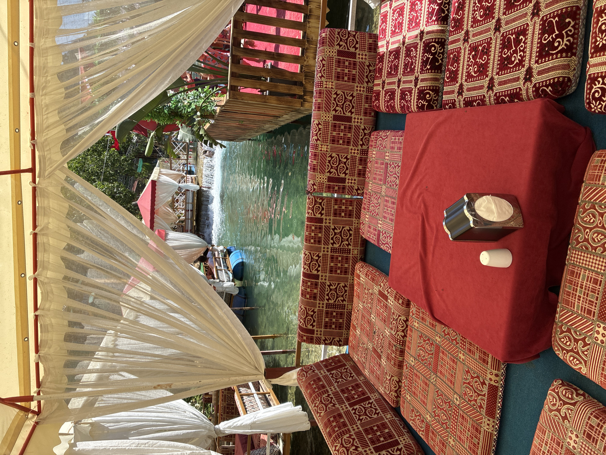 This image depicts a waterside seating area at the Dim River in Alanya, Turkey. The foreground features floor seating arranged with cushions adorned with a red and gold patterned fabric. A red tablecloth covers a low table, on which sits a metal napkin dispenser and a small white cup. Behind the seating area, a turquoise river flows, with a small waterfall visible further back. On the river, several floating platforms or boats are visible, each equipped with red-and-white canopied seating areas. The scene is partially enclosed by white sheer curtains and framed by a light-colored awning. Lush green foliage and trees are present in the background, suggesting a natural, outdoor setting.