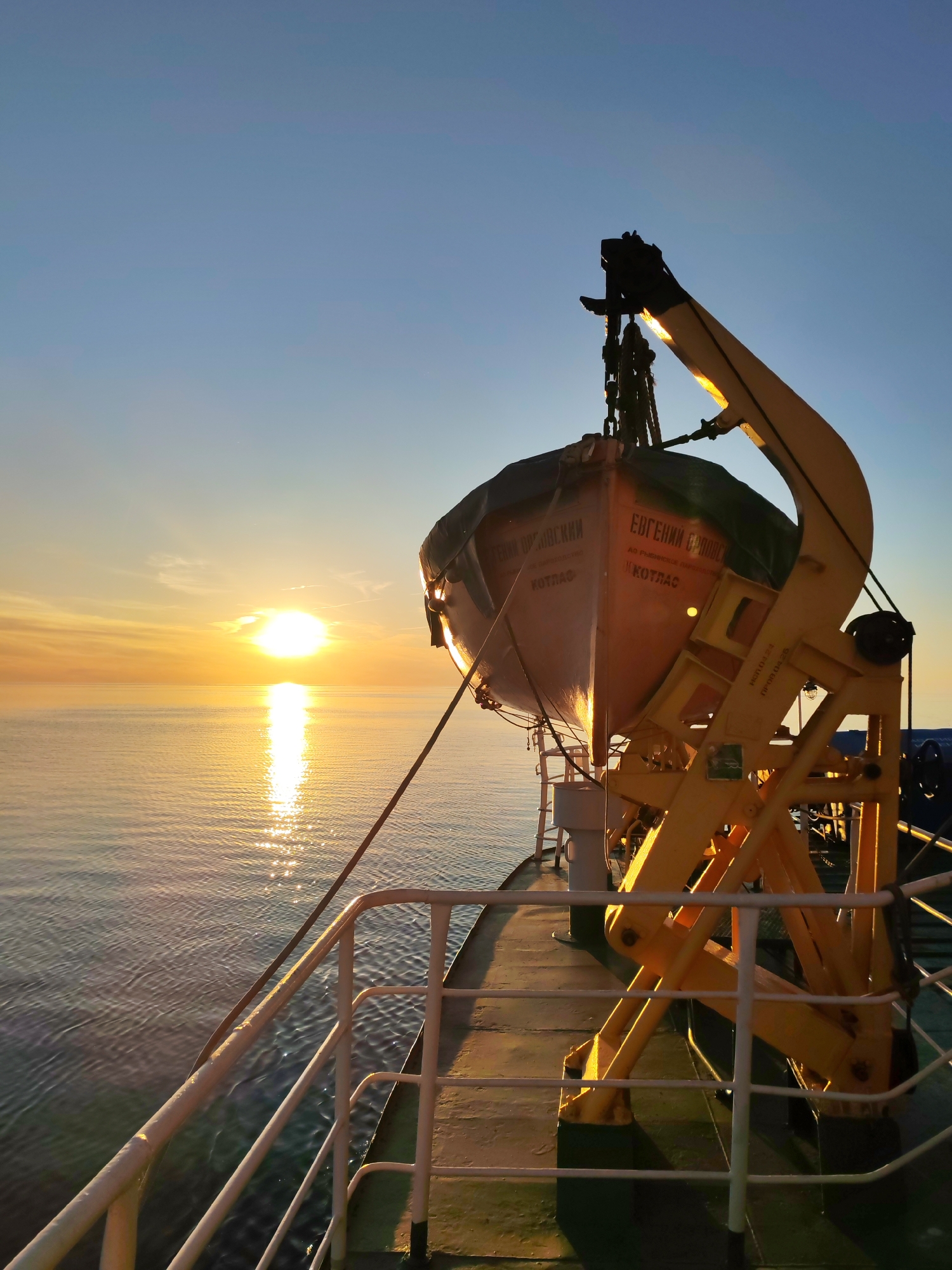 The image captures a maritime scene at sea during either sunrise or sunset, viewed from the deck of a vessel. The sun, a bright orange-yellow orb, is positioned low on the horizon on the left, casting a strong, reflective column of light across the calm water surface. The sky transitions from a light blue at the top to warm orange and yellow hues near the horizon.
In the mid-ground, a light orange, fully enclosed lifeboat is secured on a large, bright yellow davit structure on the ship's starboard side. The lifeboat has a dark covering on its upper section. Russian text is visible on the lifeboat's hull: "ЕВГЕНИЙ БРОВКИН" (Evgeniy Brovkin), "АО РЫБИНСКОЕ ПАРОВОЗНОЕ" (JSC Rybinsk Locomotive), and "КОТЛАС" (Kotlas), indicating the vessel's name, possibly its owner or builder, and its port of registration or namesake city in Russia. The yellow davit structure displays further text, "ИСП. 01.04.22" (Inspection 01.04.22) and "ПРОВ. 01.04.25" (Provision/Test 01.04.25), likely indicating dates of inspection and re-provisioning.
The ship's deck, visible in the foreground and lower right, is a dark grey or green, with white railings extending along the side. A dark rope descends from the lifeboat towards the deck. The low sun angle creates strong highlights on the surfaces of the davit and lifeboat facing the sun, while other areas are in shadow or silhouette. No people are visible in the frame. The location is at sea; there is no discernible cityscape.
